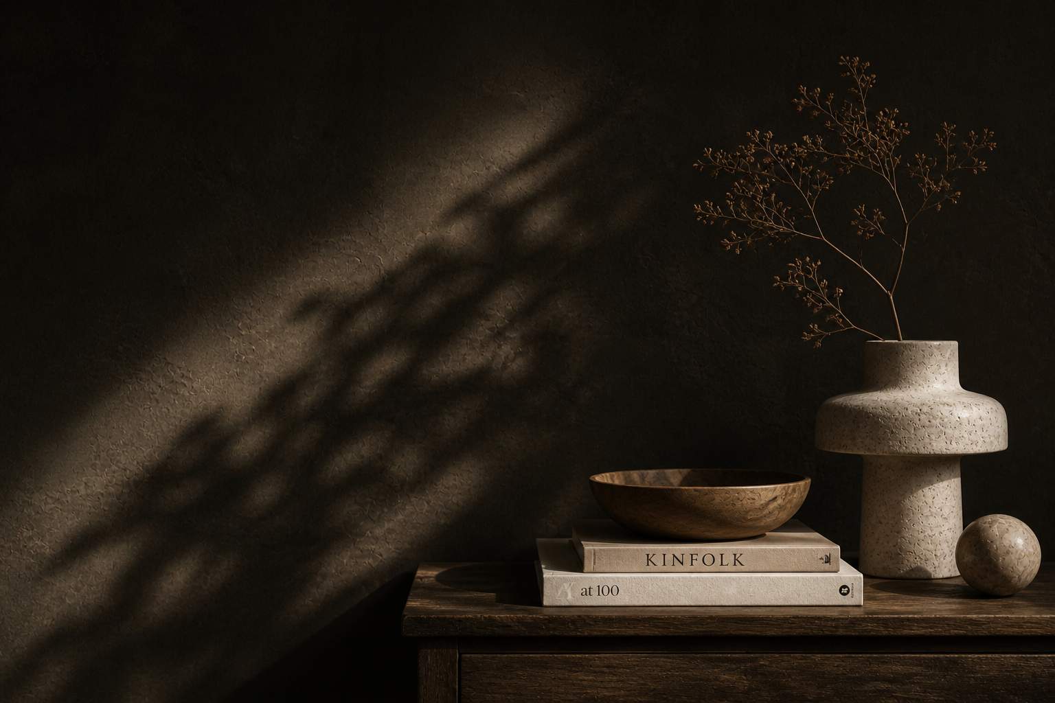Decorative arrangement on a dark wooden surface including a textured ceramic vase with a single dried branch, a round stone object, a wooden bowl, and books titled "Kinfolk" and "at 100" with a shadow cast by the branch on the dark wall behind.
