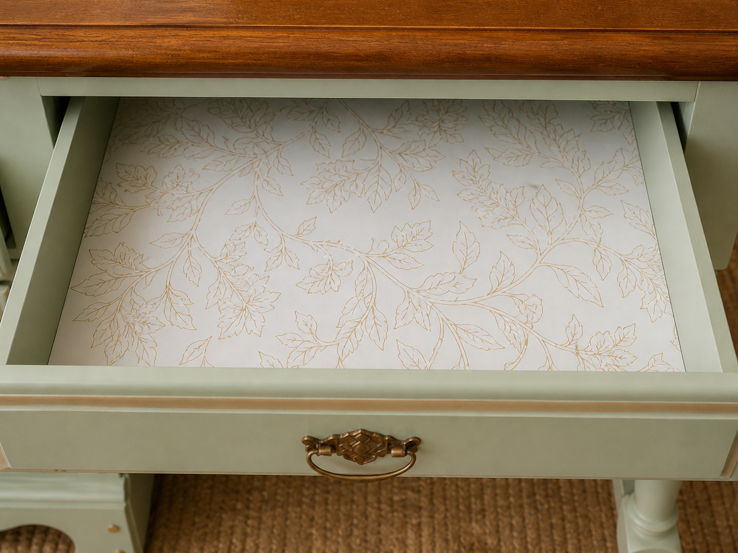 Open drawer with floral patterned paper lining, beige leaves on a white background, wooden drawer handle, part of a light-colored furniture piece, and a brown carpeted floor.