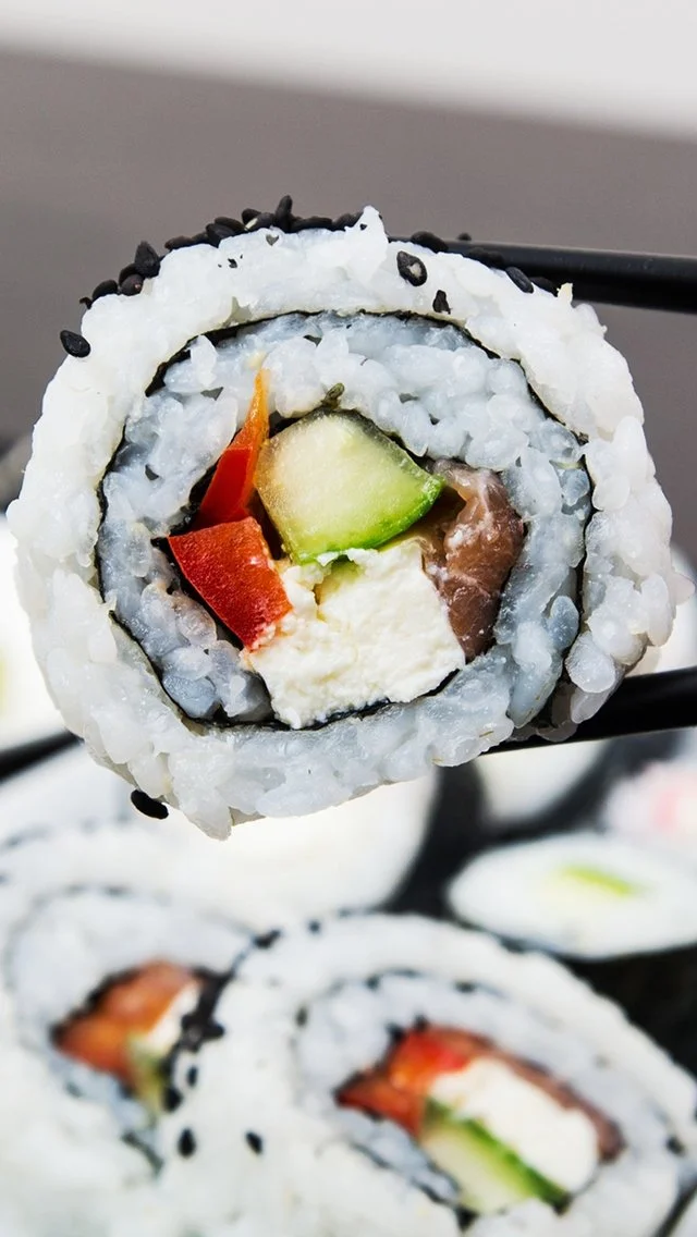 Close-up of a sushi roll filled with cucumber, red bell pepper, cream cheese, and fish, topped with rice and black sesame seeds.