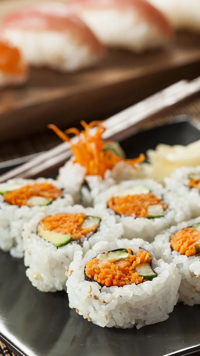 Close-up of sushi rolls with rice, carrots, and cucumber on a black plate, with pickled ginger in the background.