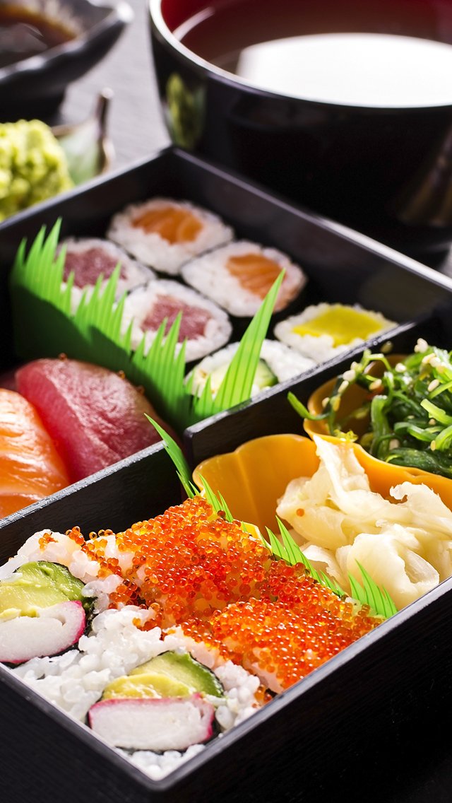 A bento box containing sushi rolls with fish, vegetables, and fish roe, accompanied by slices of raw fish, cucumber, and pickled ginger, with a bowl of miso soup in the background.