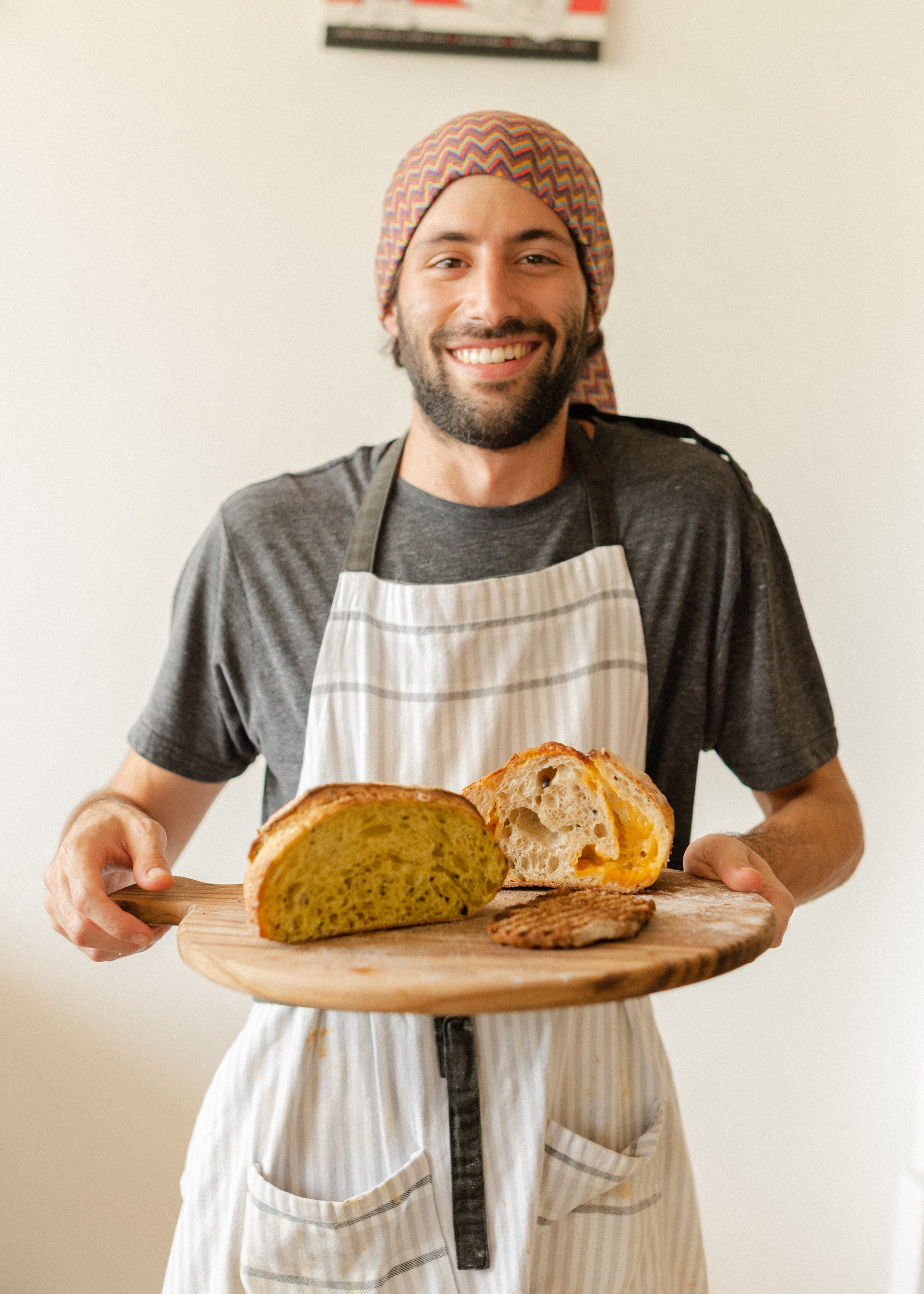Hombre con gorro y delantal sostiene una tabla con panes y carne, sonriendo.