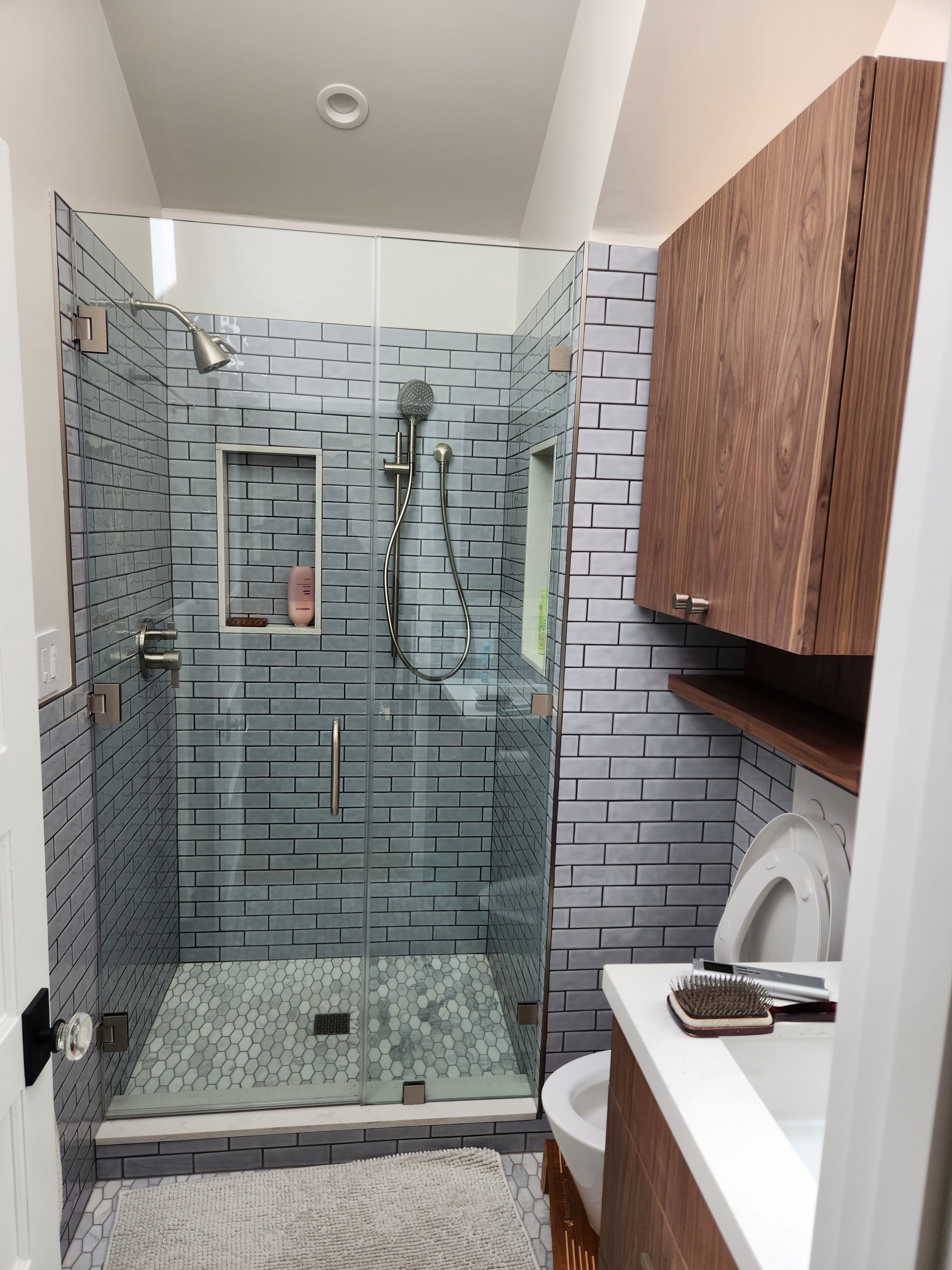A modern bathroom with a glass-enclosed walk-in shower, gray subway tiles, a wooden cabinet, a toilet, and a white countertop with grooming tools.