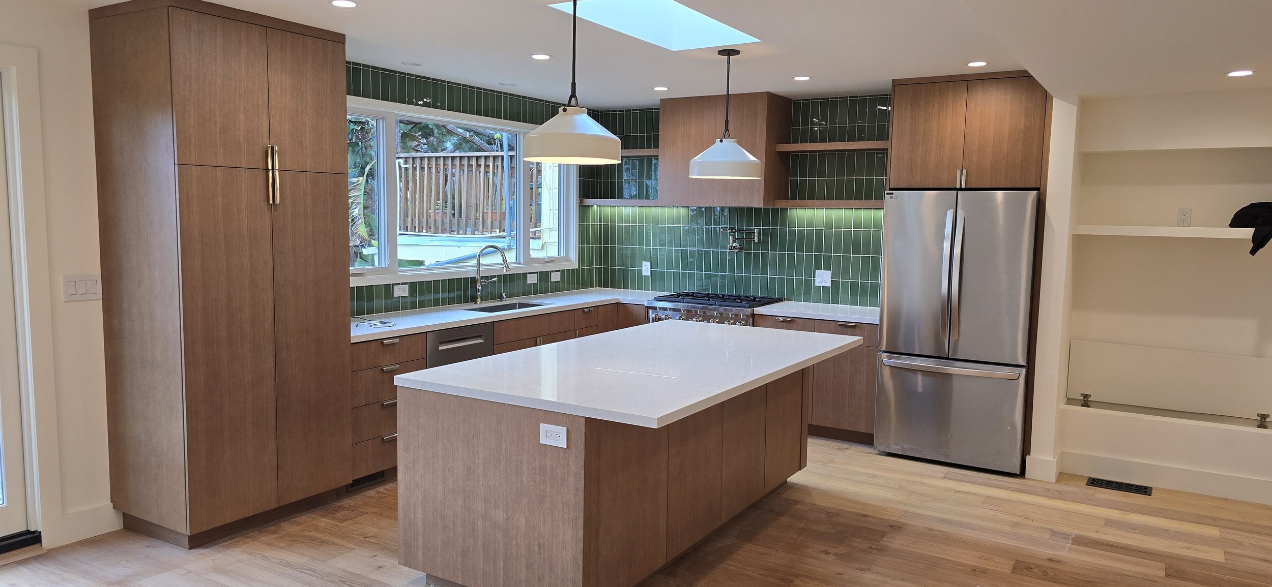 Modern kitchen with wooden cabinets, green tile backsplash, white countertop island, stainless steel appliances, and a large window overlooking a backyard.