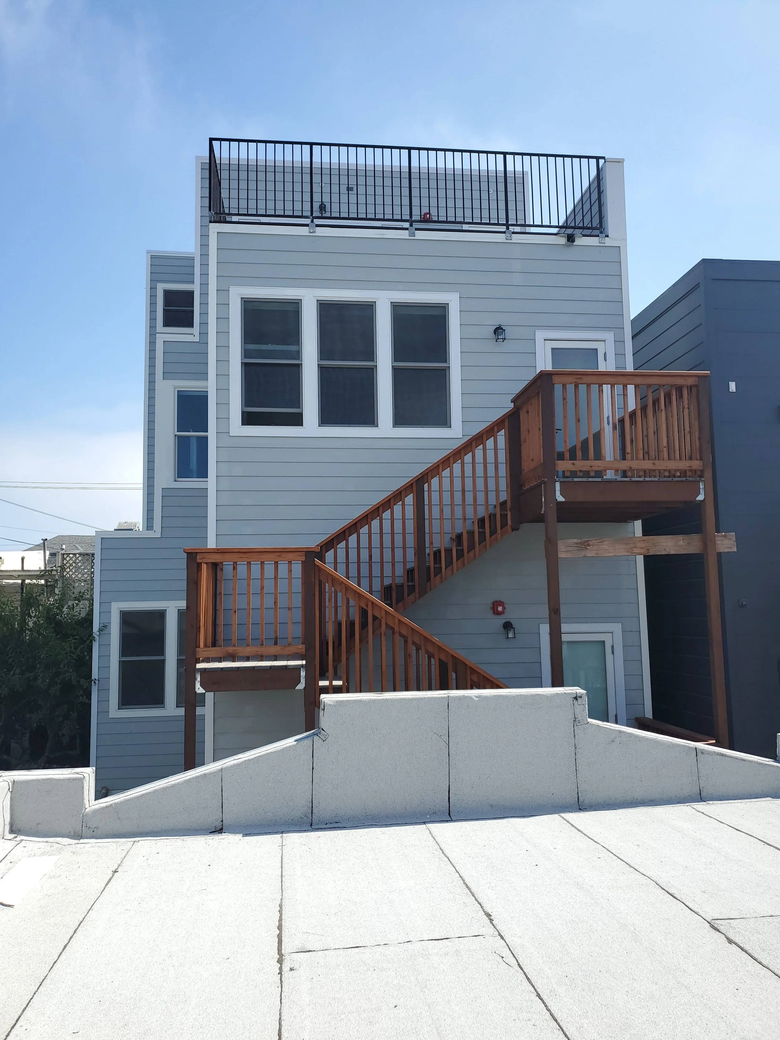 Multi-story residential building with exterior staircase, multiple windows, gray siding, and a rooftop balcony with black railing, under a blue sky.