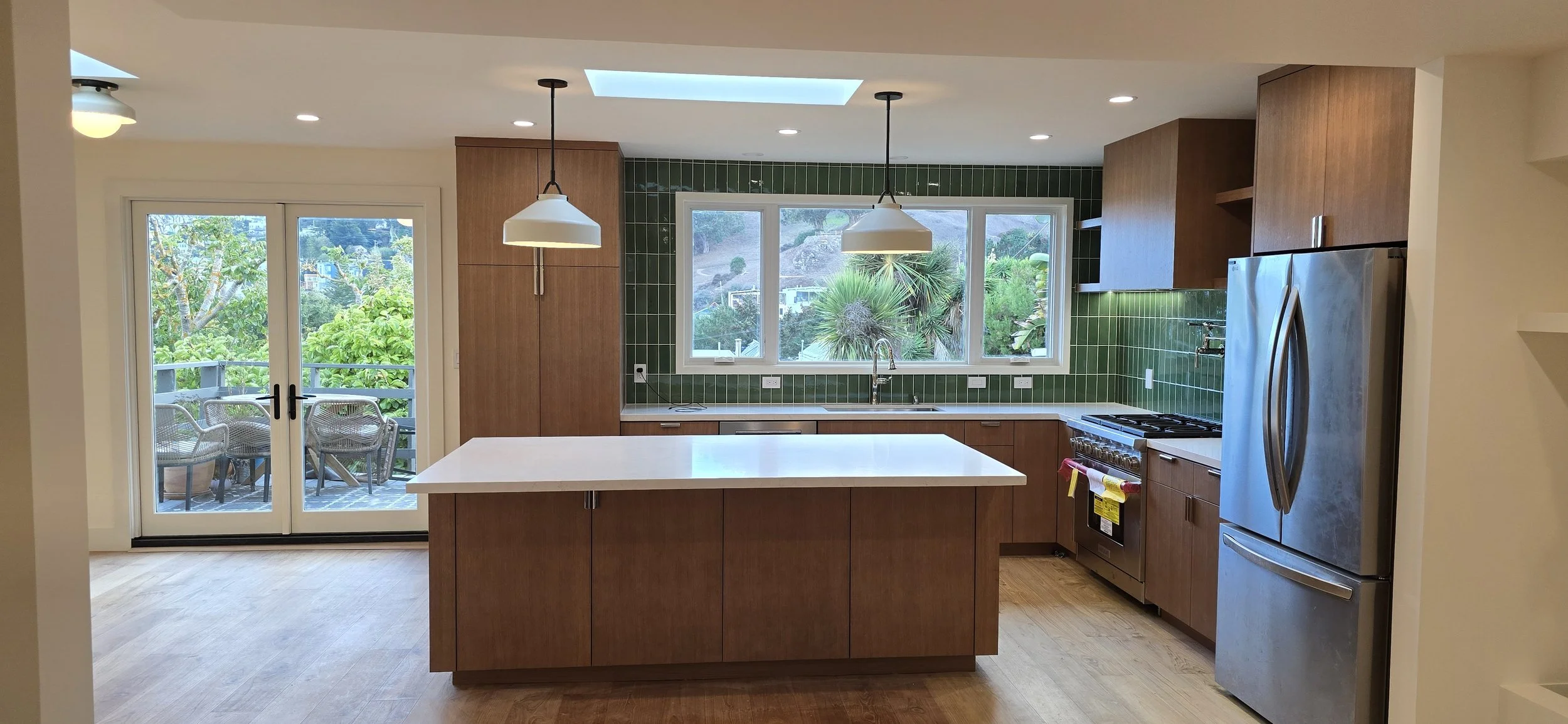 Modern kitchen with green tiled backsplash, wooden cabinets, white countertop island, stainless steel appliances, large window showing outdoor greenery, and glass sliding door leading to patio with outdoor furniture.