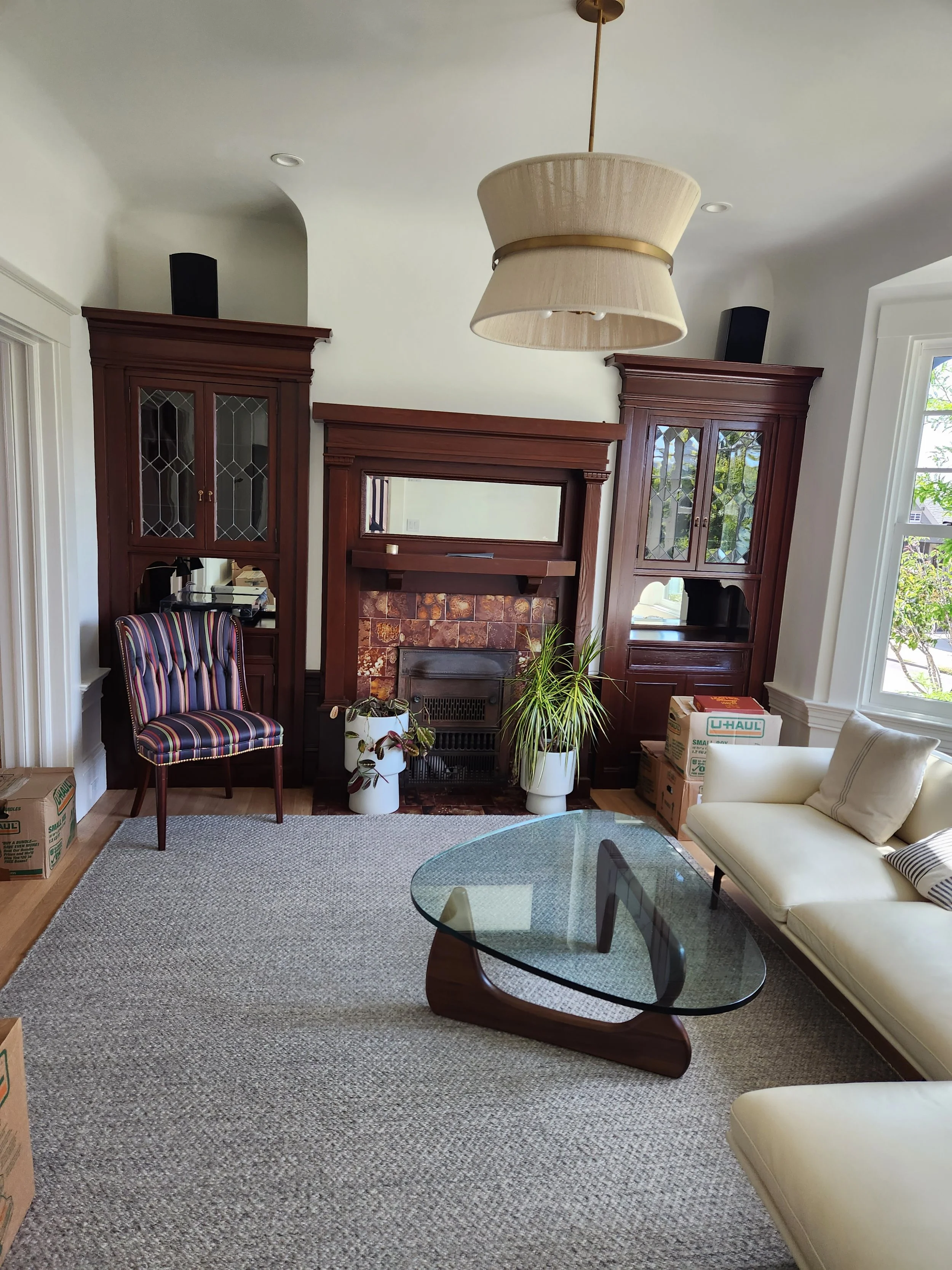 Living room with cream-colored sofa, glass coffee table, fireplace with wooden mantel, built-in wooden cabinets, and moving boxes near the window.