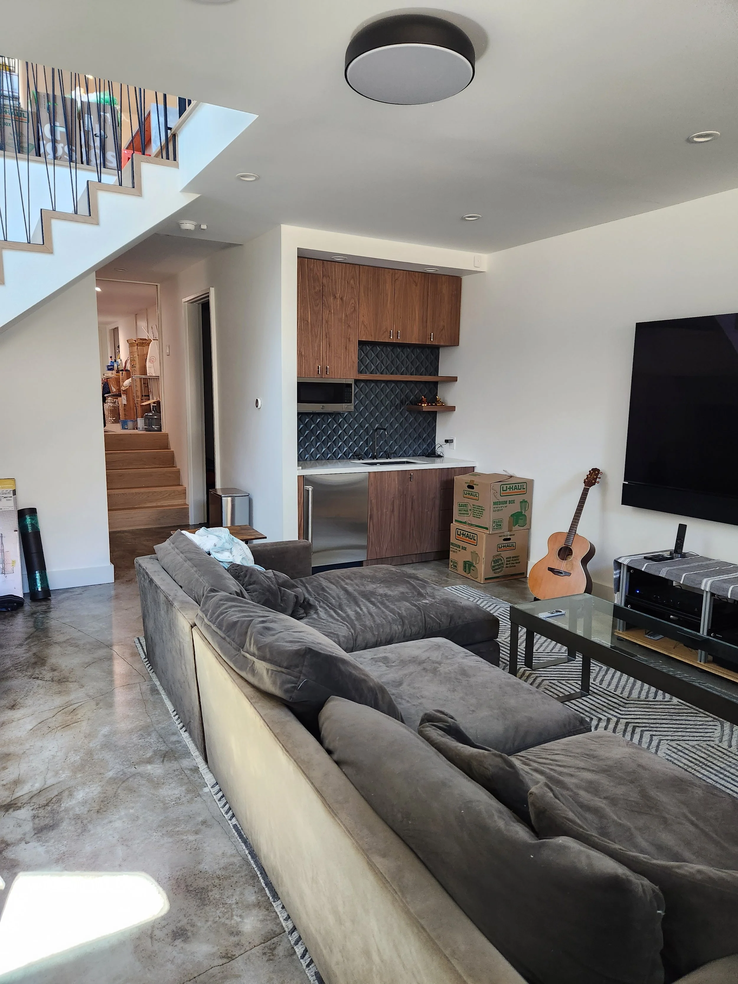 Living room with large sectional sofa, coffee table, mounted TV, guitar, and boxes. Open kitchen area with wooden cabinets and black tile backsplash. Staircase on left leads to upper level.