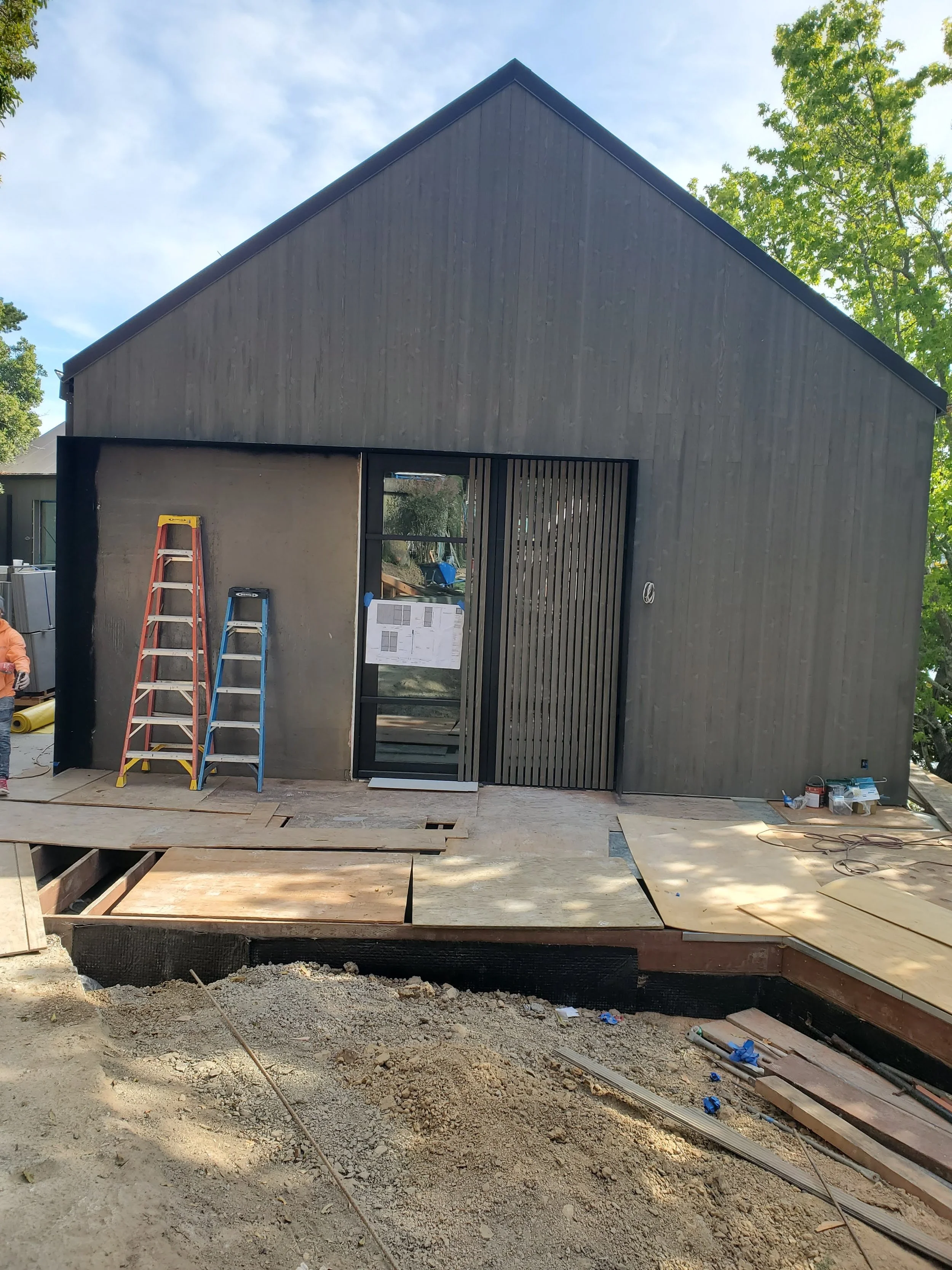 A black house under construction with ladders, work supplies, and wooden planks on the porch, and a partially built front yard with dirt and construction debris.