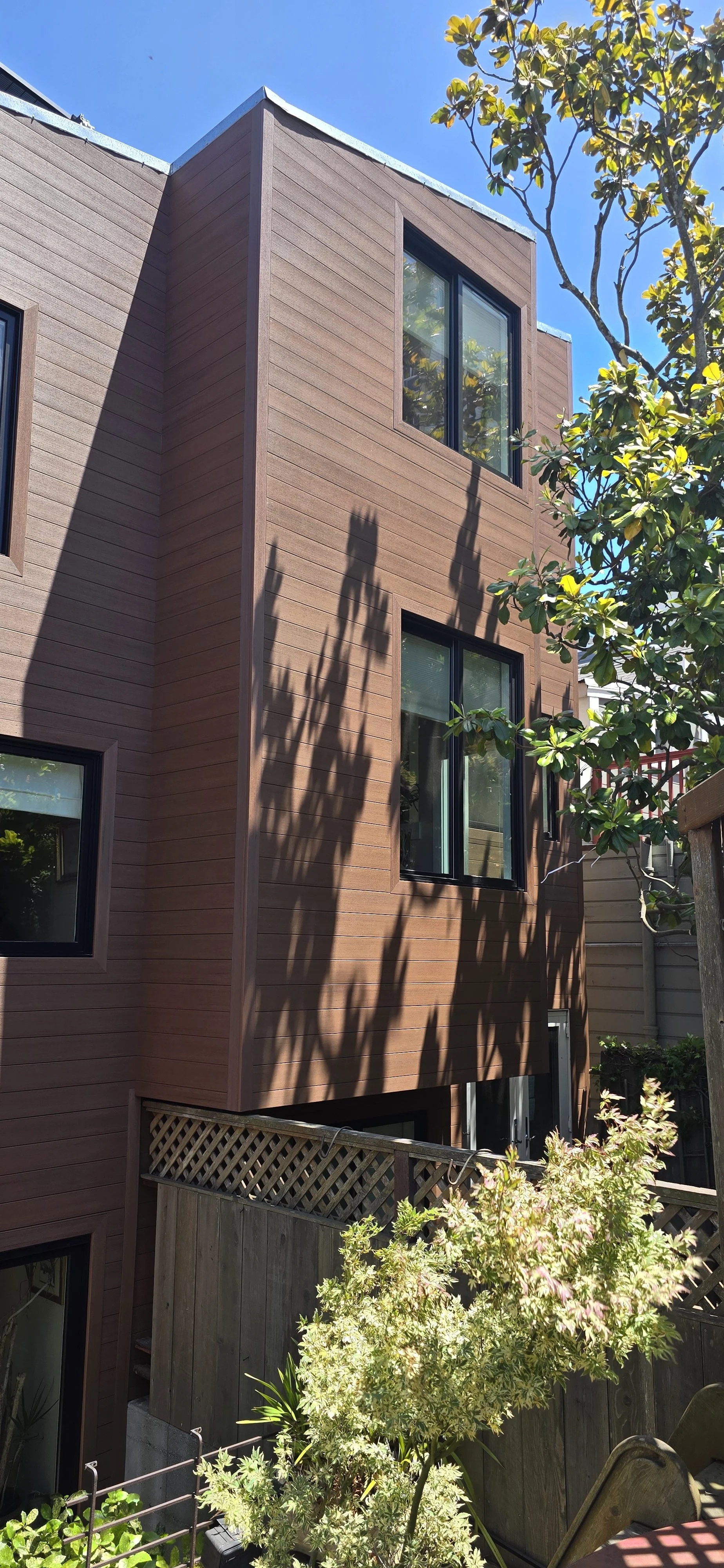 A modern multi-story house with brown wooden siding, black-framed windows, and a small fenced yard with plants and trees. Shadows of the trees fall on the house's exterior.