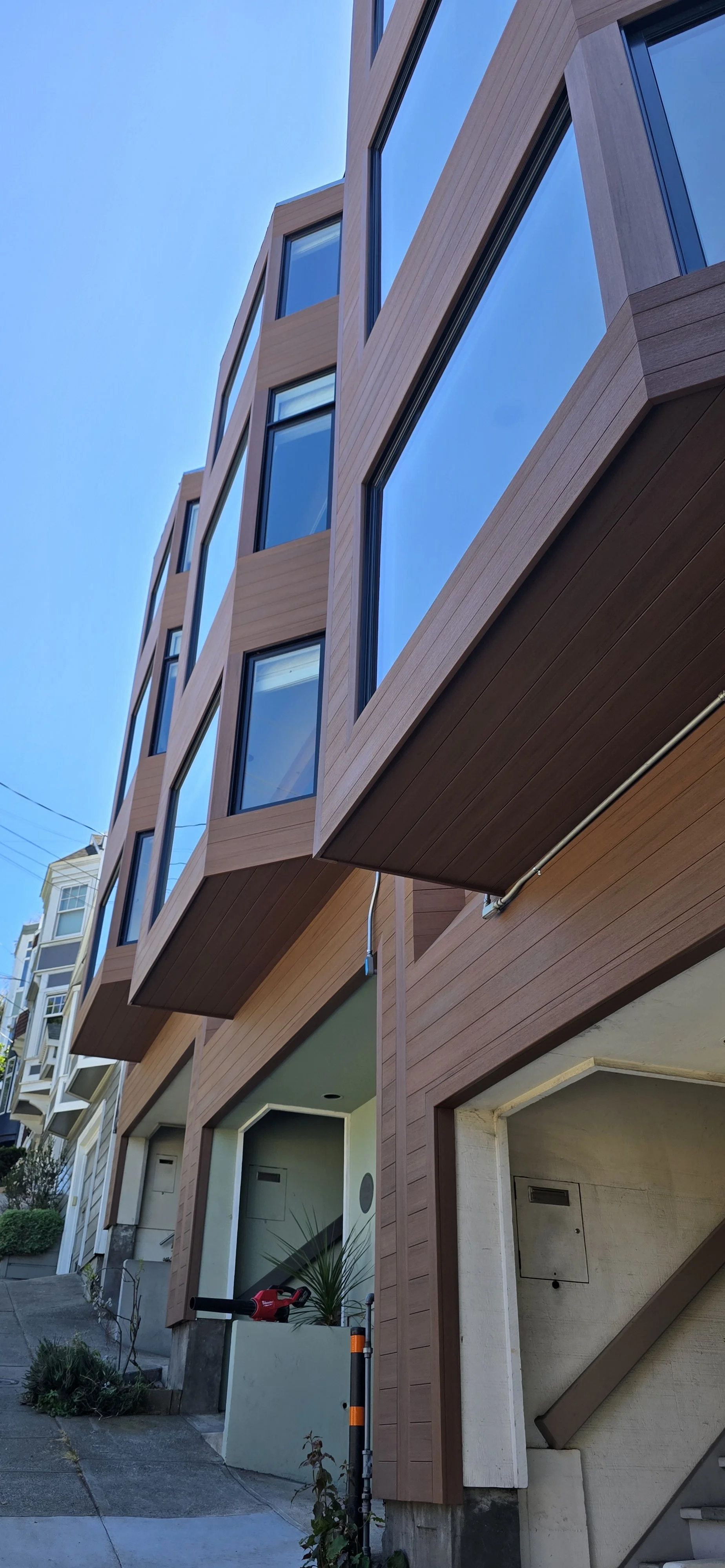 Modern multi-story building with large windows and wooden exterior panels, situated on a sloped street under a clear blue sky.