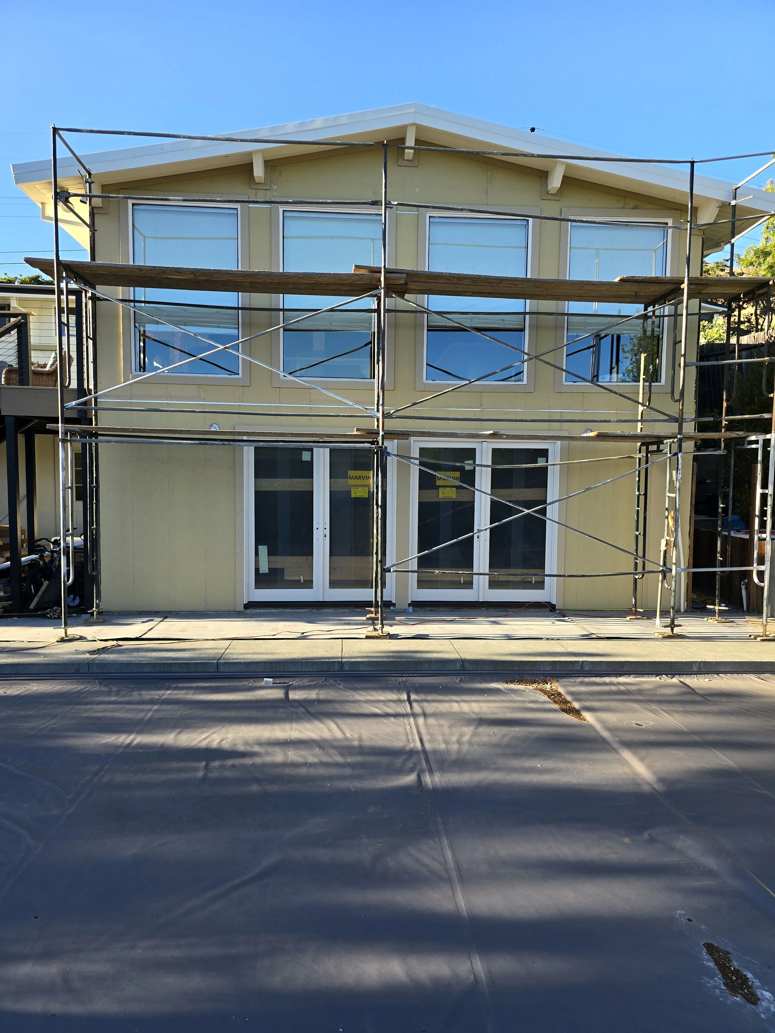 Two-story house under construction with yellow siding, large windows, and scaffolding surrounding the house. The house has a sloped roof and is situated on a paved street with shadows cast on the ground.