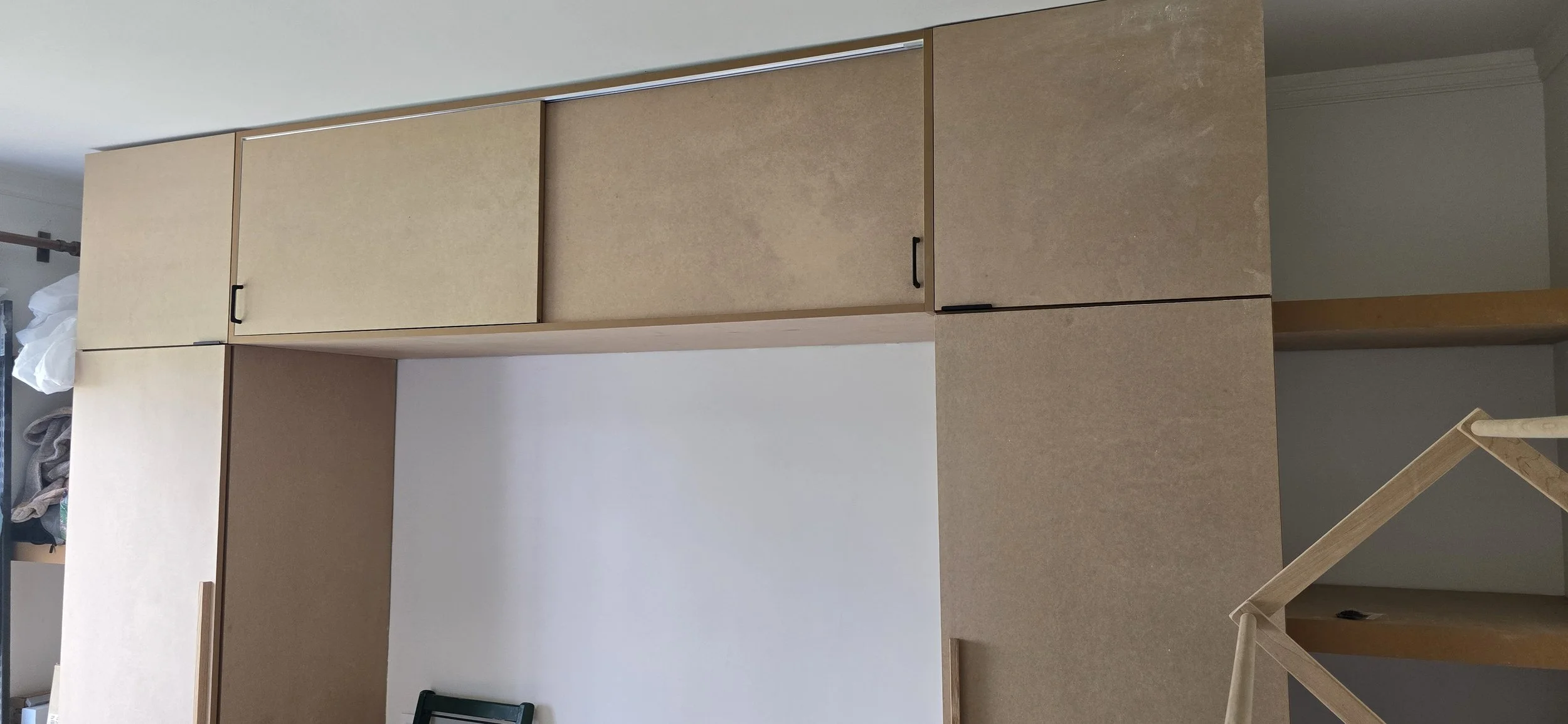 Image of a partially assembled wooden built-in wardrobe with multiple compartments, doors, and black handles, situated in a room with white walls and crown molding.