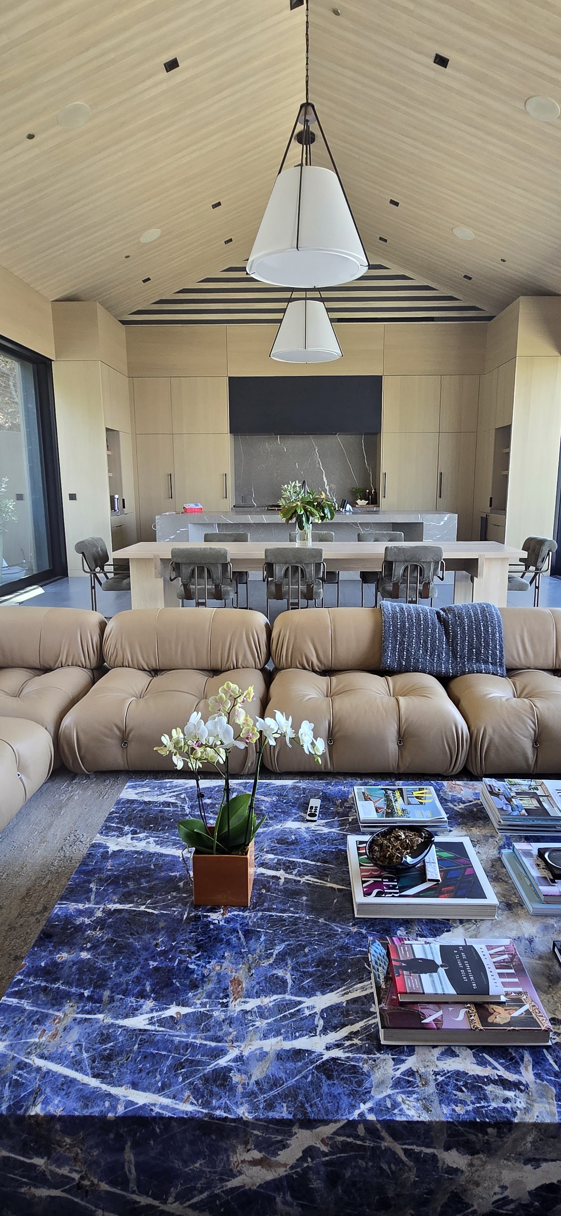 Modern living room with beige leather sectional sofa, blue marble coffee table, and a vase of white orchids. In the background, an open kitchen with light wood cabinets, a marble island, and a dining table with eight upholstered chairs. Large sliding