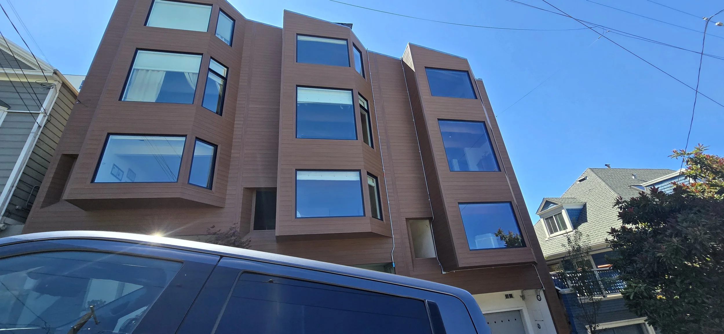 Multi-story residential building with a brown facade and large windows, situated in a neighborhood with other houses, clear blue sky above, and a black vehicle partially visible in the foreground.