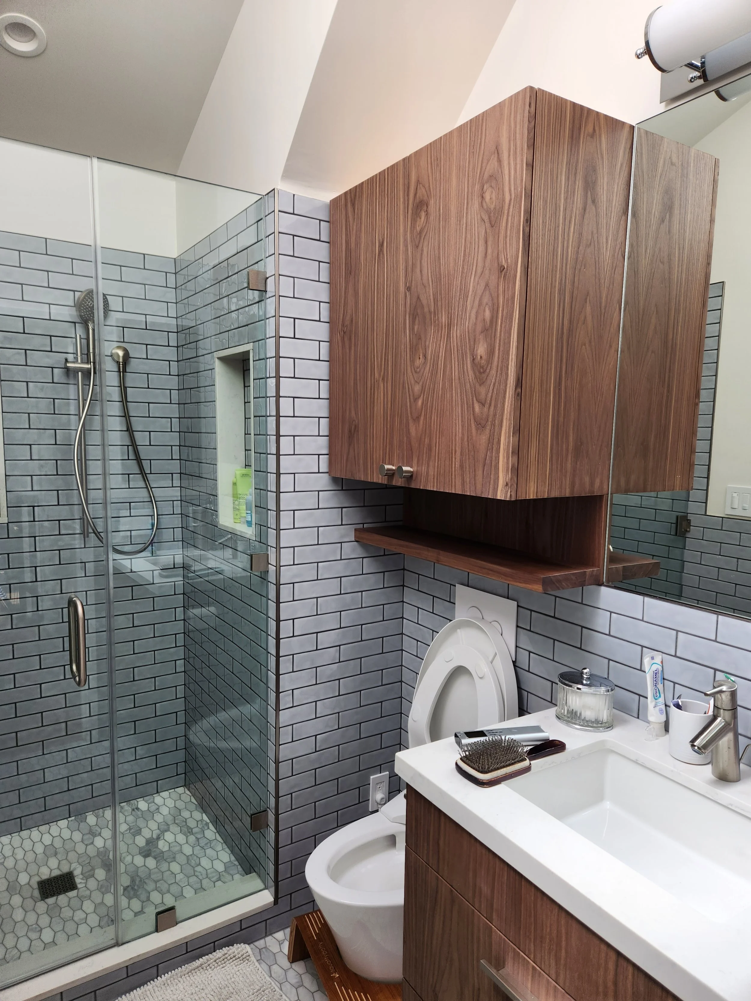 Modern bathroom with a glass shower, wooden cabinet above the toilet, and a white sink with bathroom essentials on the countertop.