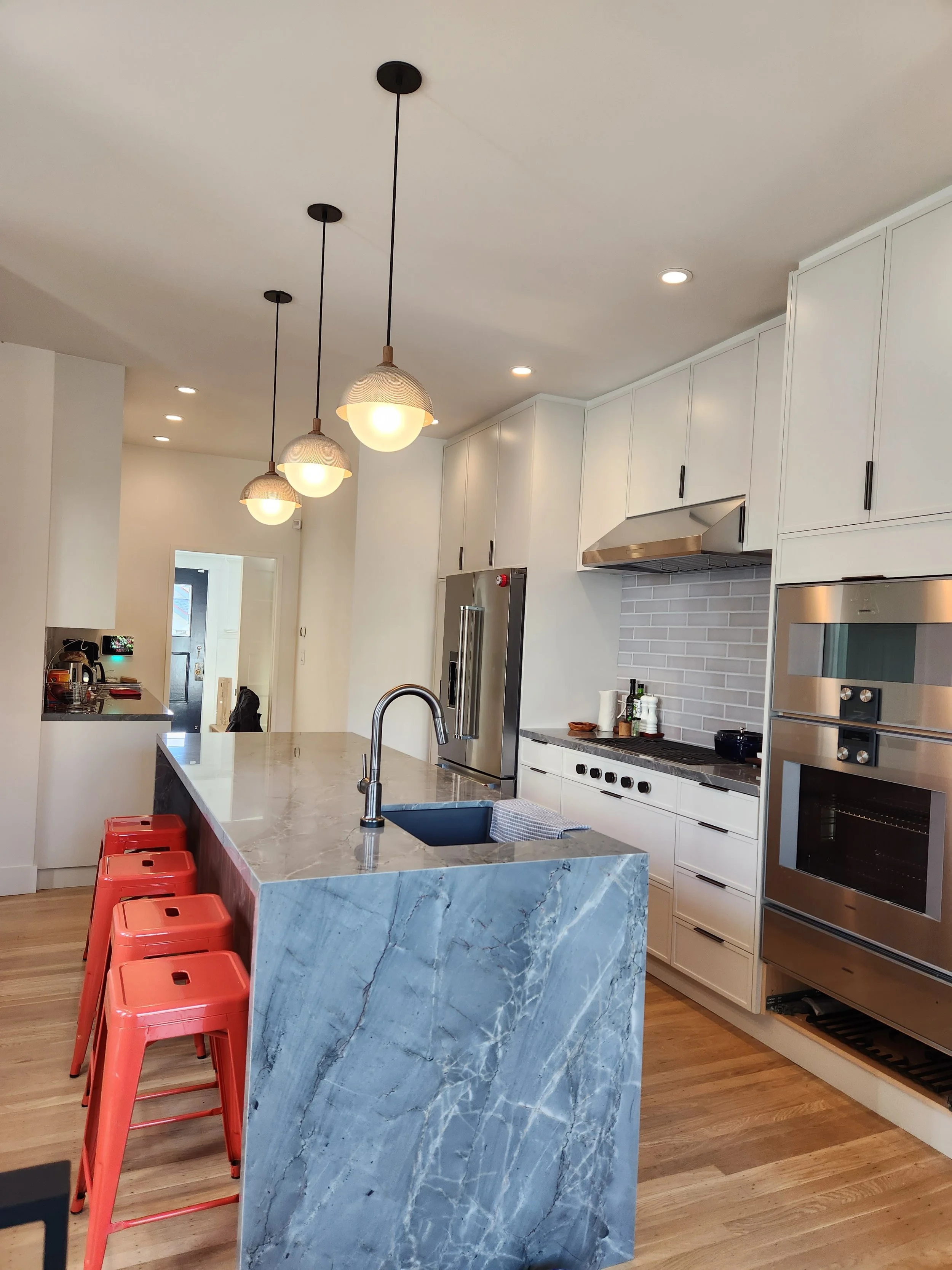 Modern kitchen with white cabinets, stainless steel appliances, a marble island with a sink, four red barstools, pendant lights, and hardwood flooring.