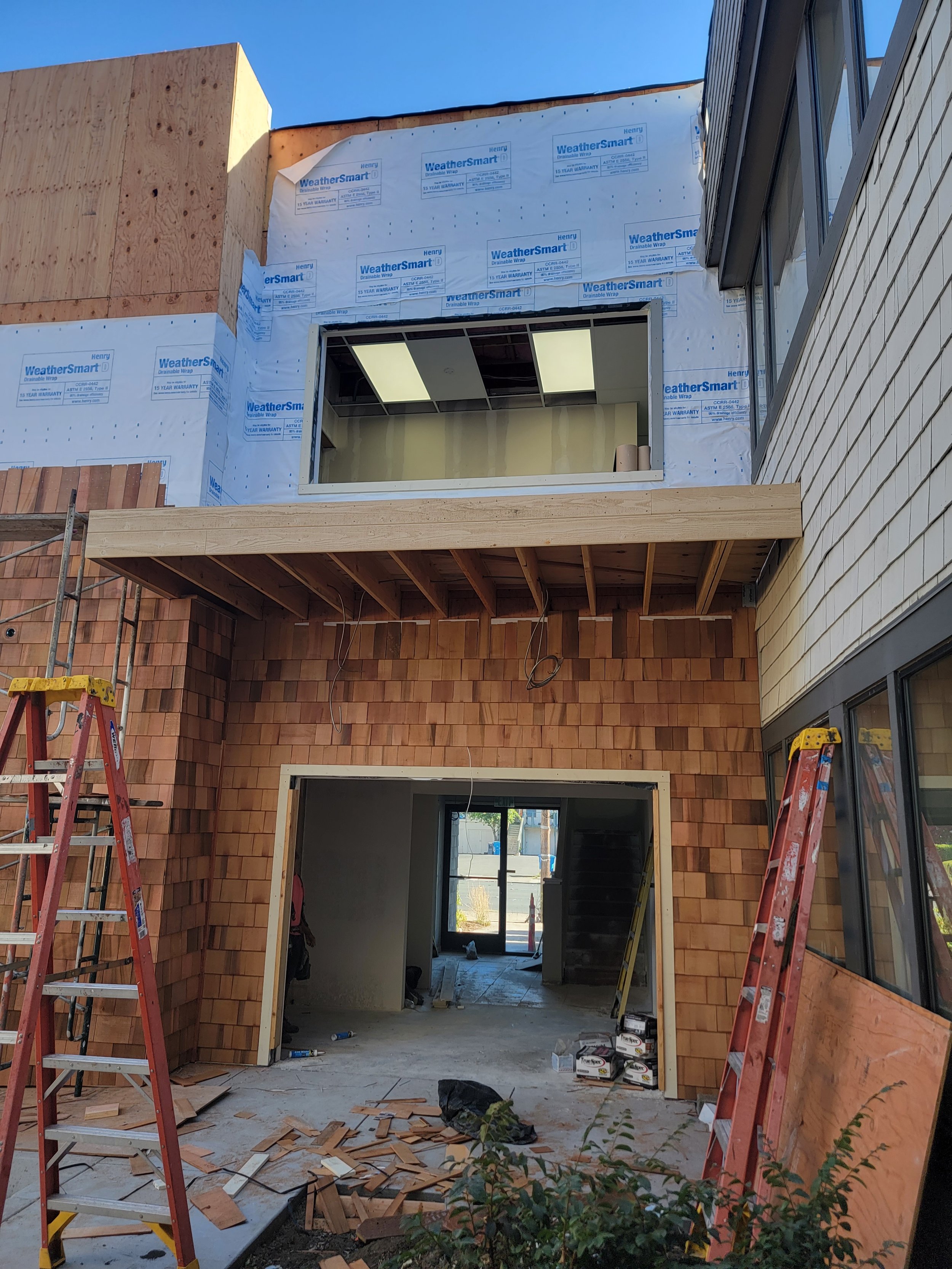 Construction site showing a building with an open garage entrance, unfinished brick exterior, scaffolding, ladders, building materials, and a large window with ceiling lights inside.