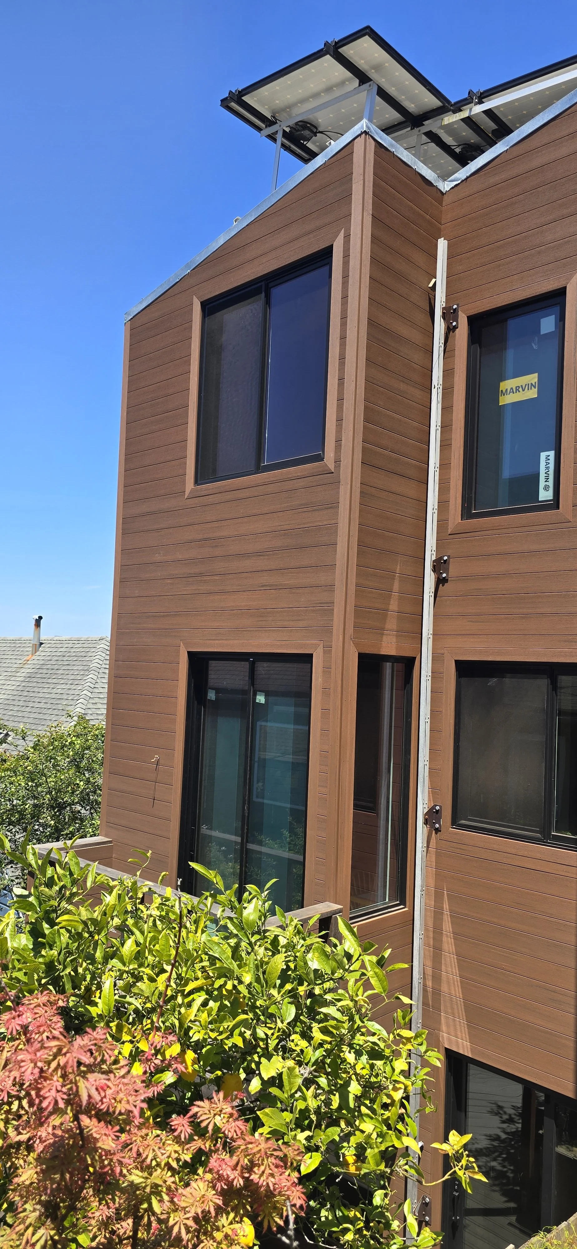 A modern multi-story building with wood siding, large black-framed windows, and solar panels on the roof. There are green and reddish plants in the foreground.