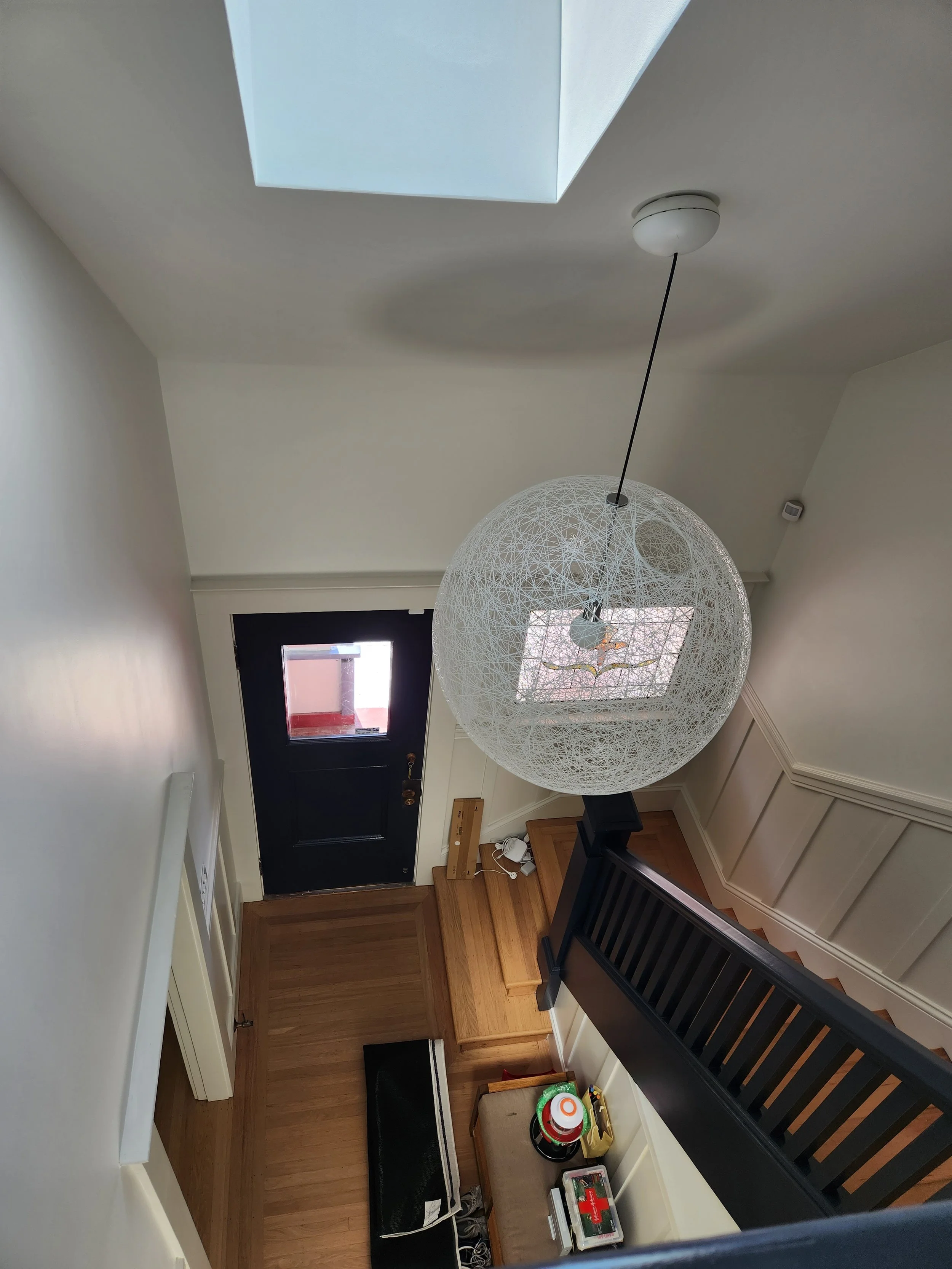 Interior view of a house entryway with a black front door, wooden flooring, a staircase with black railing, a large decorative spherical white pendant light, and a skylight in the ceiling.