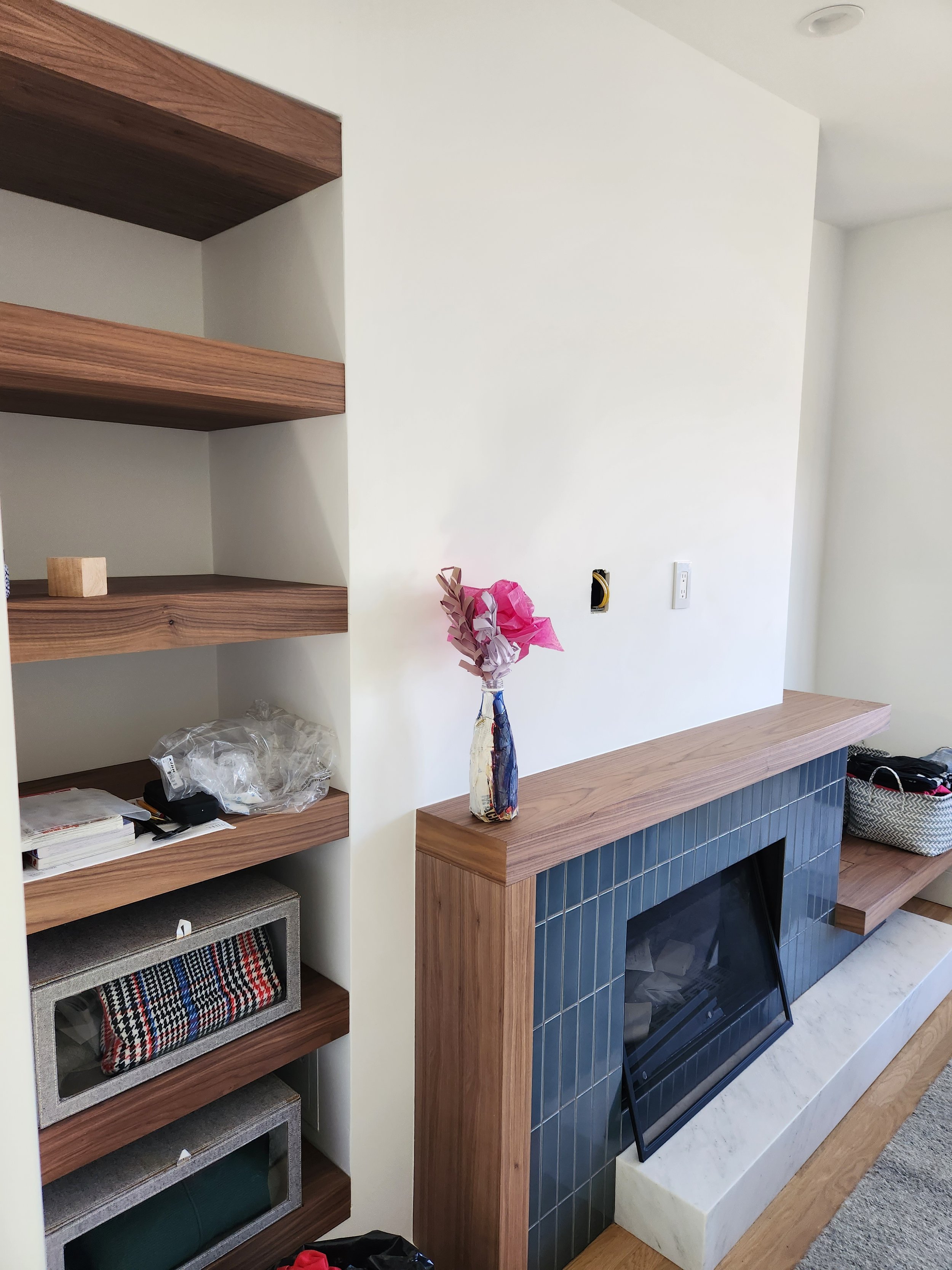 Interior of a living room featuring a wooden and tiled fireplace with a glass cover, a dark blue tiled border, a floating wooden shelf, a decorative bottle with pink tissue paper flowers, and open shelving with miscellaneous items.
