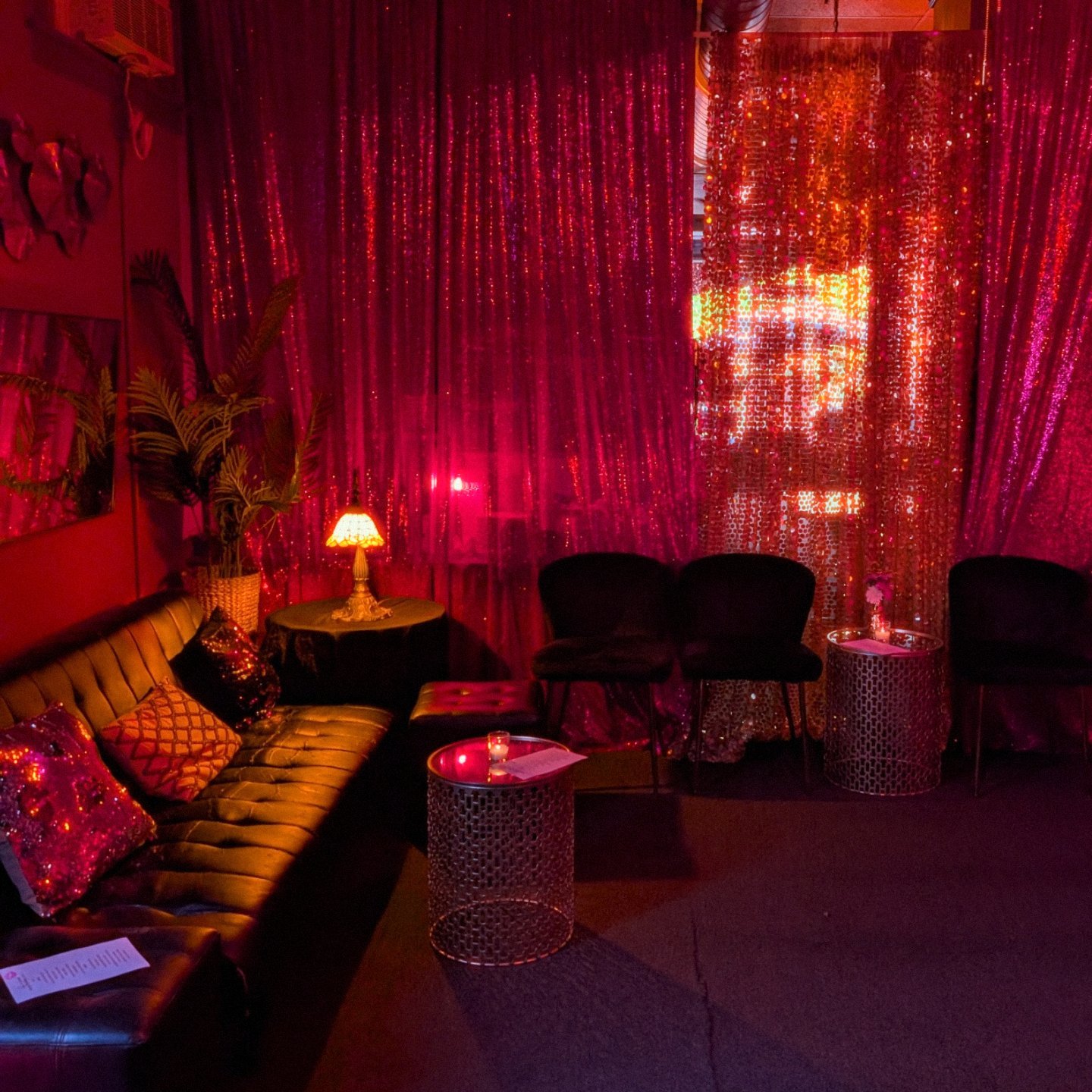 Dimly lit room with pink and red lighting, a sofa with colorful pillows, small tables with candles, black chairs, pink curtains with shiny embellishments, a small table with a lamp, and decorative plants.