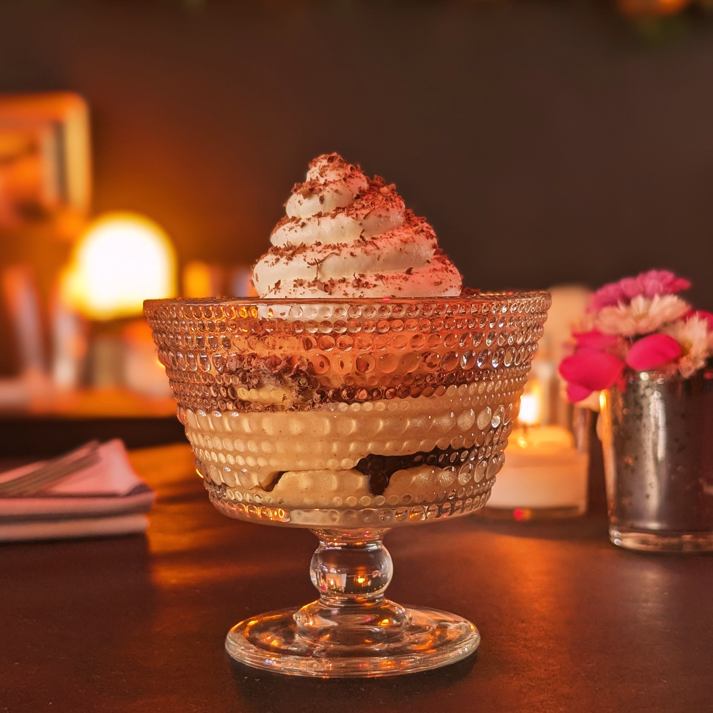 A dessert in a glass bowl topped with whipped cream and cocoa powder, served on a dark table with a blurred background, pink flowers in a metal vase, and warm lighting.