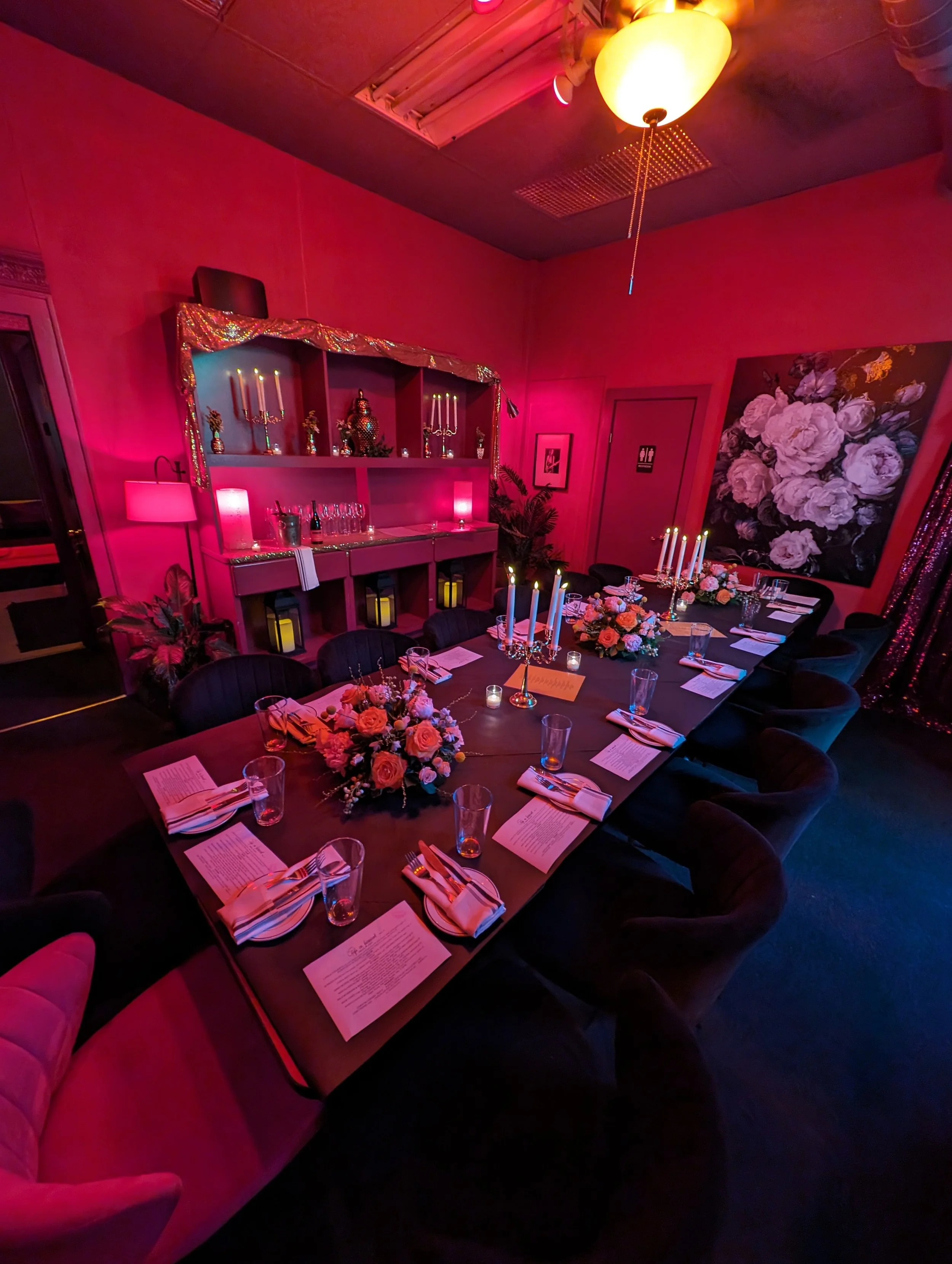 A decorated dining room with pink and purple lighting, featuring a long table set with floral centerpieces, candles, notes, glasses, and wrapped napkins. The room has pink walls, a floral painting, and a cabinet with candles and decorative items.