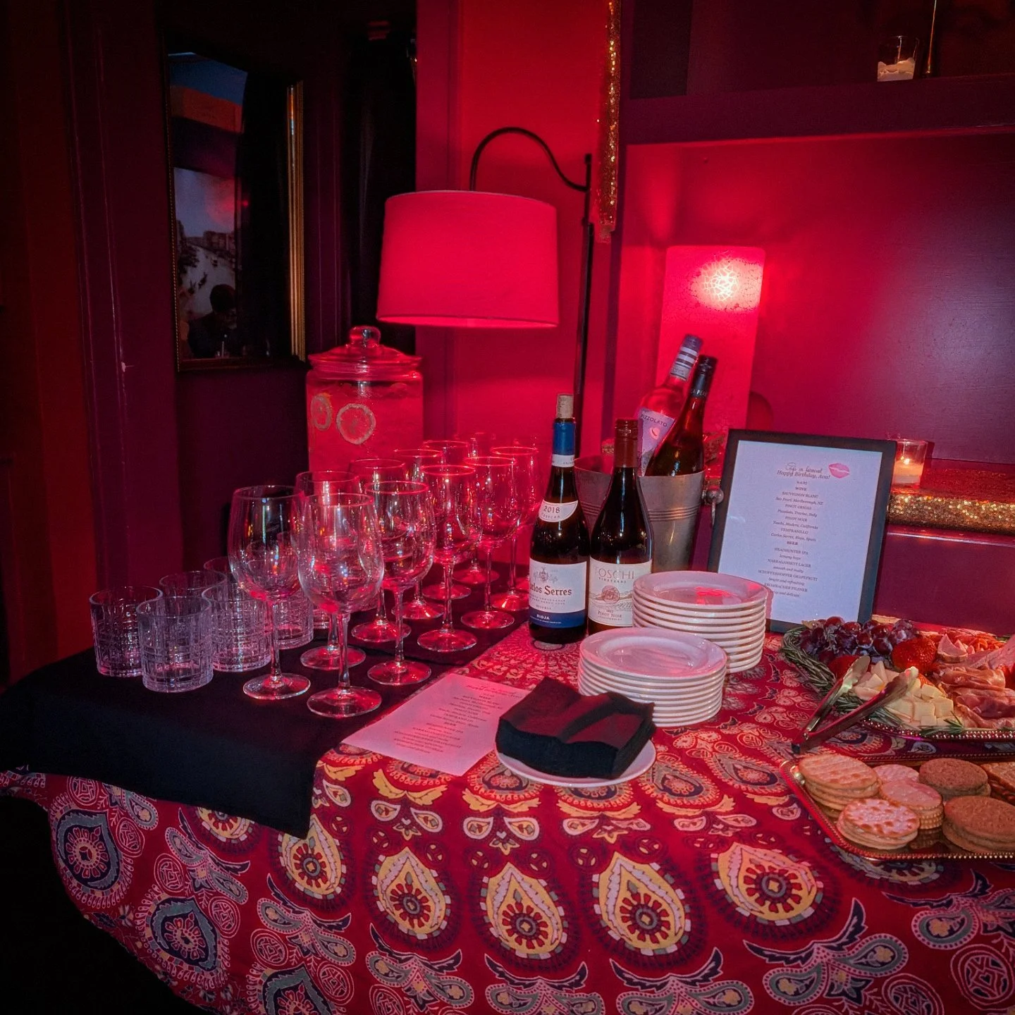 A table set with empty wine glasses, bottles of wine, a lit pink lamp, plates, a tray of assorted snacks and chocolates, and a framed menu, in a dimly lit room with pink lighting.