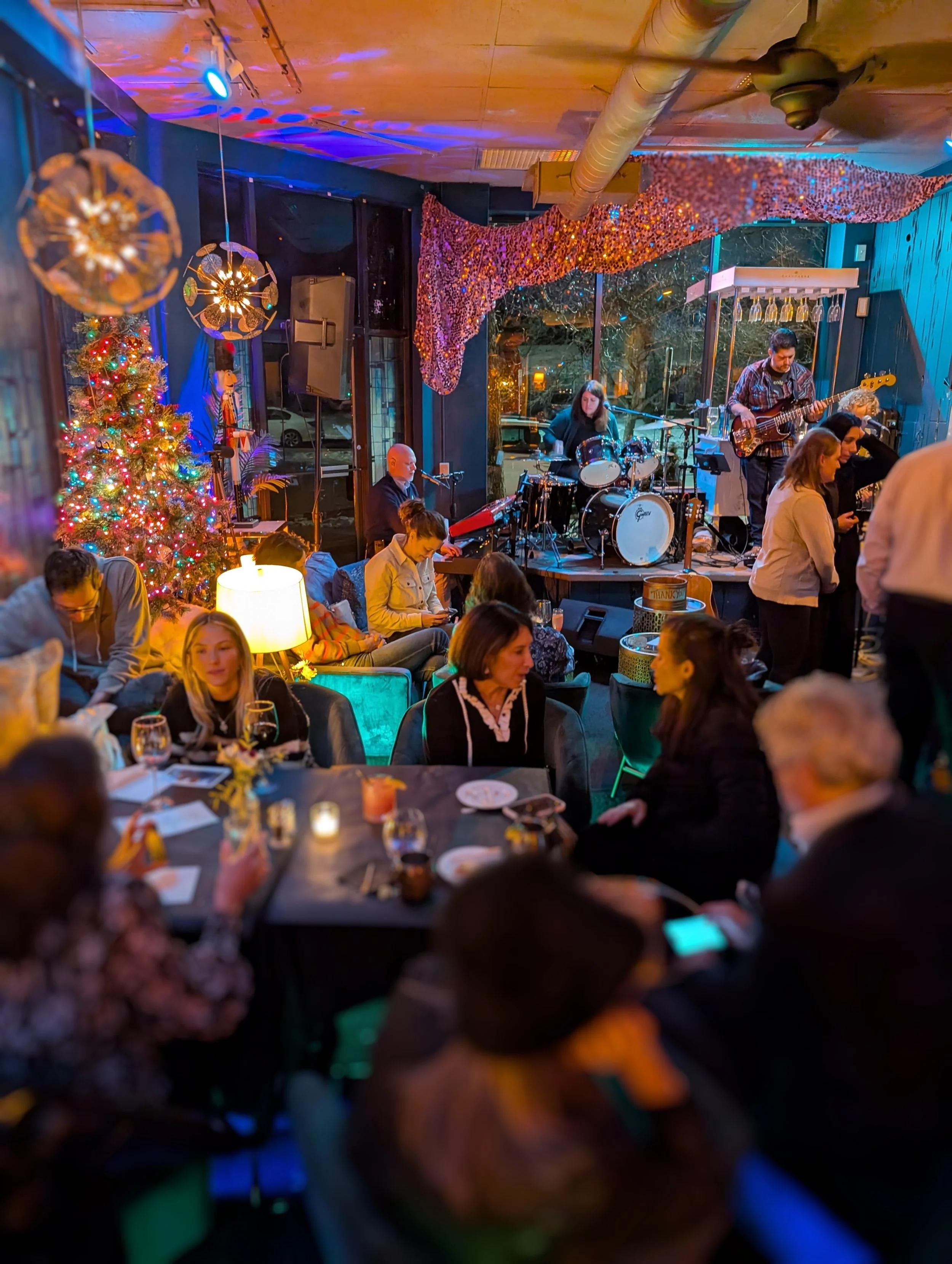 Indoor scene at a live music venue decorated for Christmas, featuring a Christmas tree with colorful lights and ornaments, a band performing on stage with drums, guitars, and keyboards, and an audience seated at tables with drinks, enjoying the atmos
