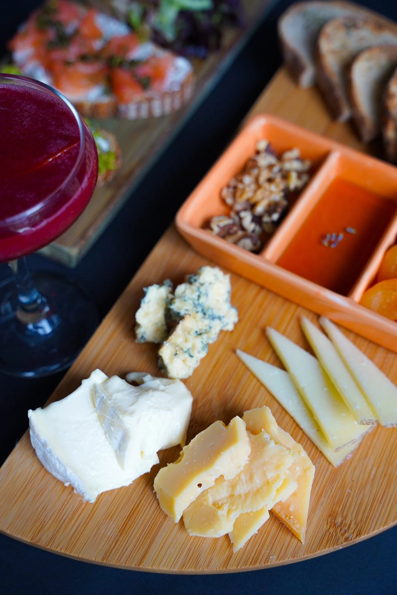 Cheese platter with various cheeses, nuts, and sliced bread, alongside a red drink, on a wooden serving board.