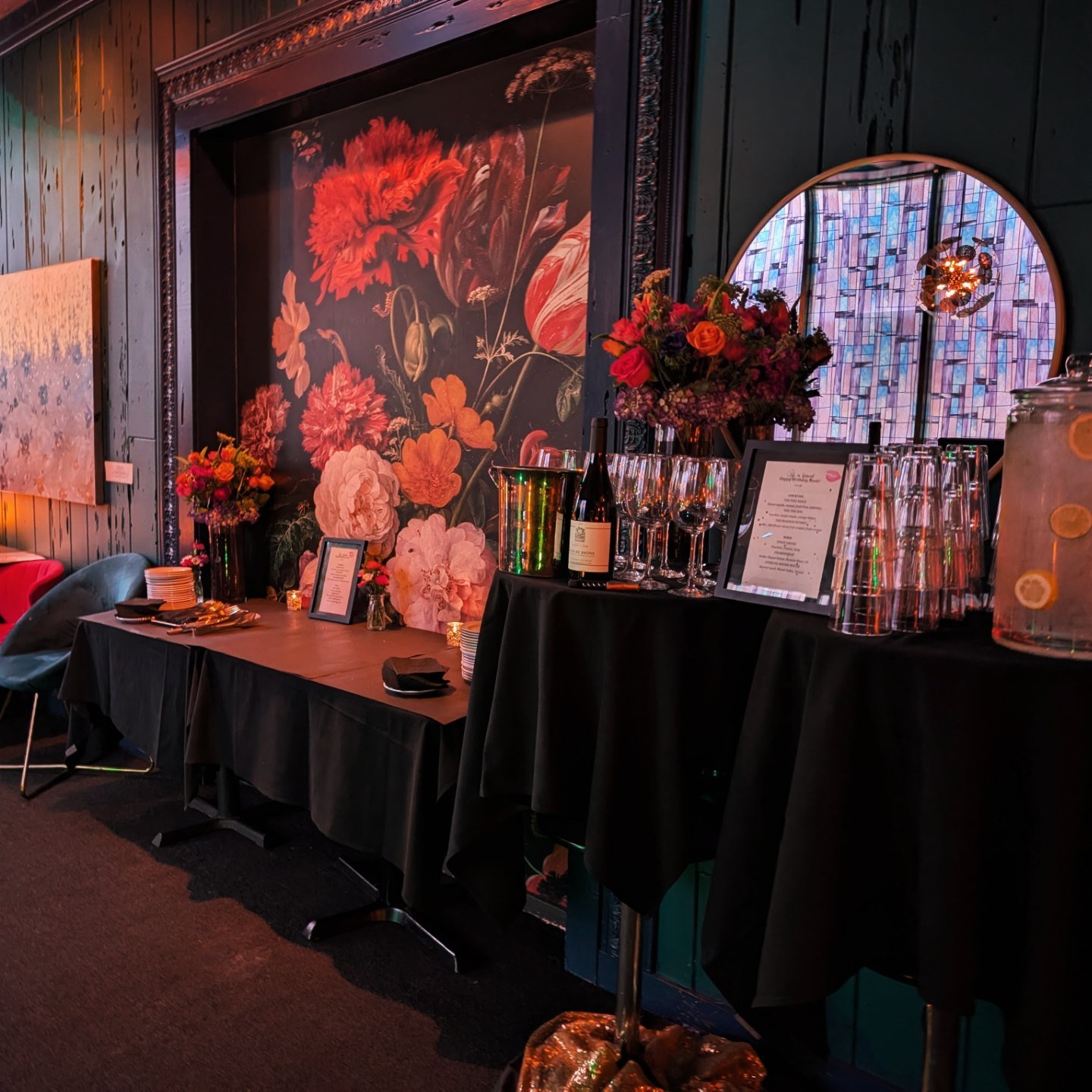 A decorated table with flowers, wine bottles, and glasses inside a dimly lit venue with dark walls and a large floral artwork on the wall.