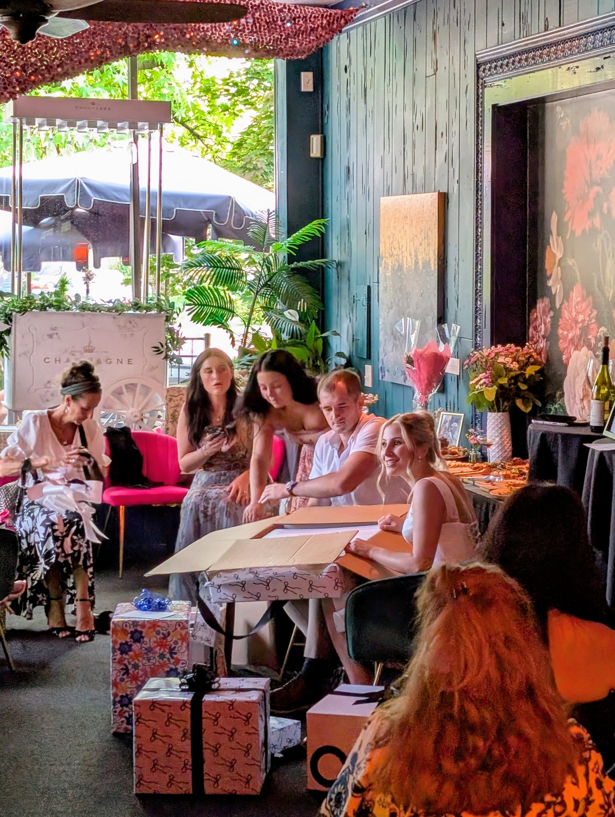 A group of people celebrating a birthday or special occasion indoors with gifts, flowers, and decorations, near an open window with outdoor greenery.