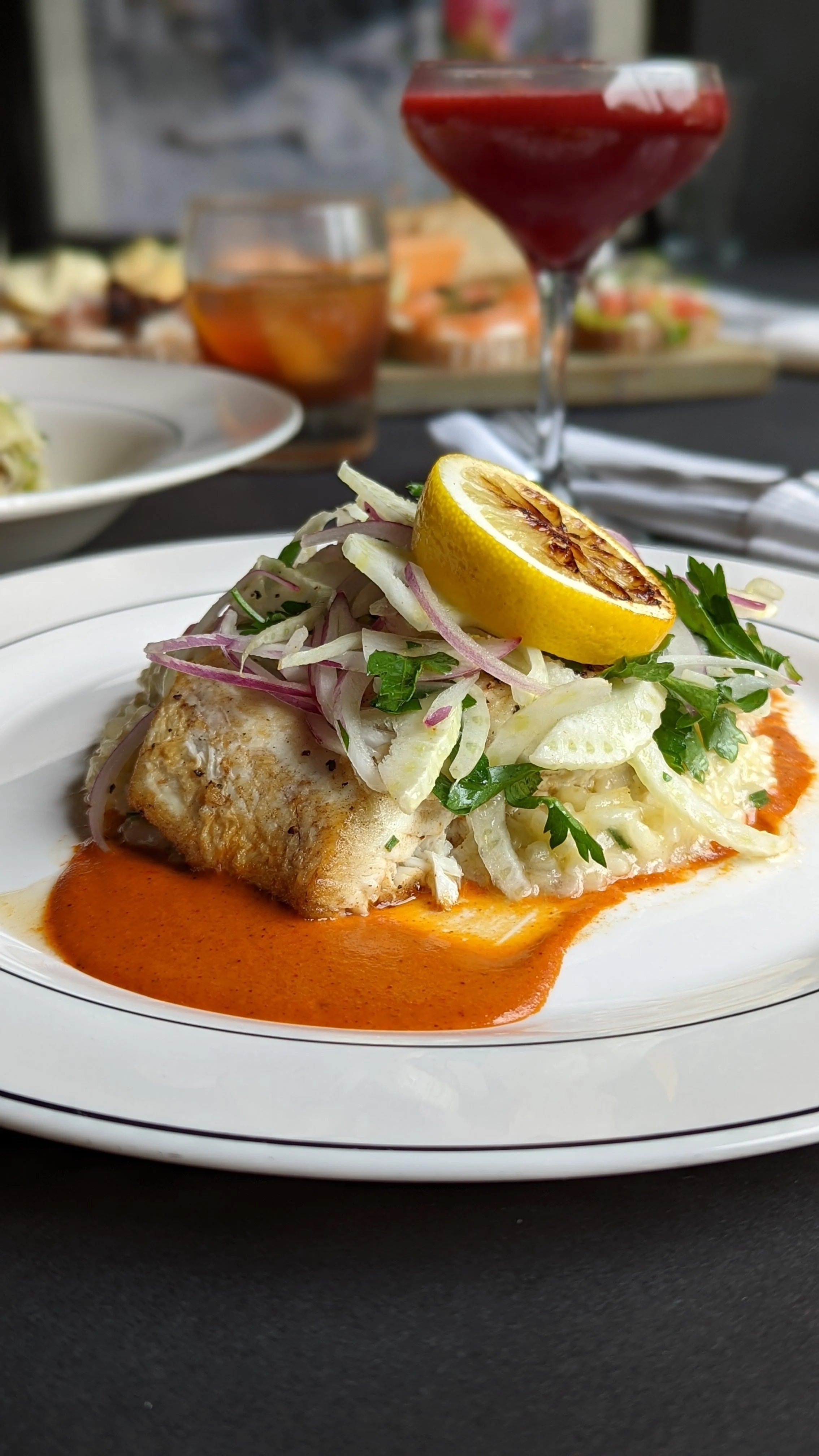 A plated serving of fish with a lemon wedge and fresh herbs on top, accompanied by a creamy sauce and a red sauce underneath. In the background, a glass of red wine, a glass of iced tea, and other dishes are visible.