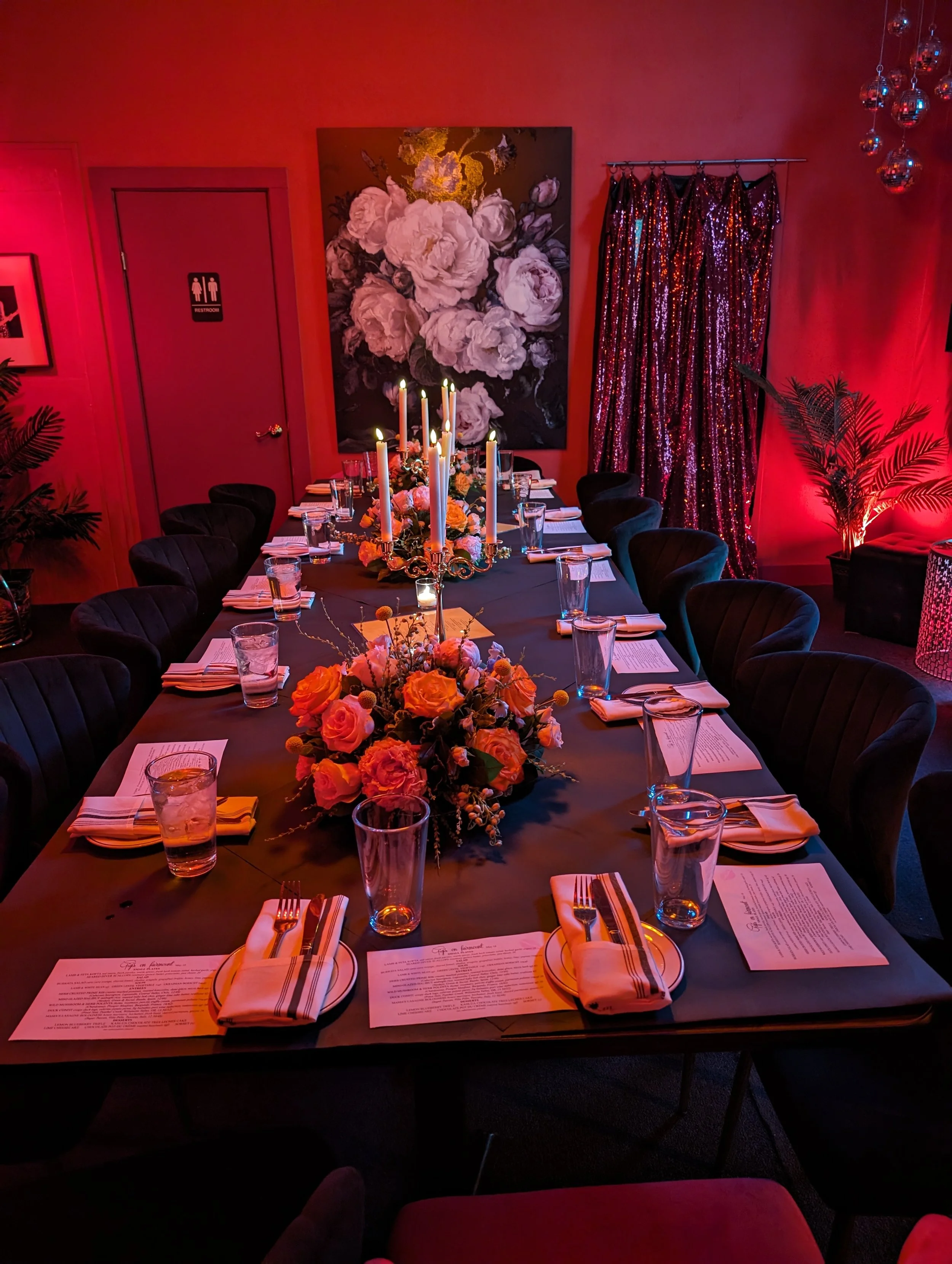 A long dining table set for a formal dinner with floral centerpieces, candles, and place settings, in a dimly lit room with red lighting and decorative elements, including a large floral painting and shimmering curtains.