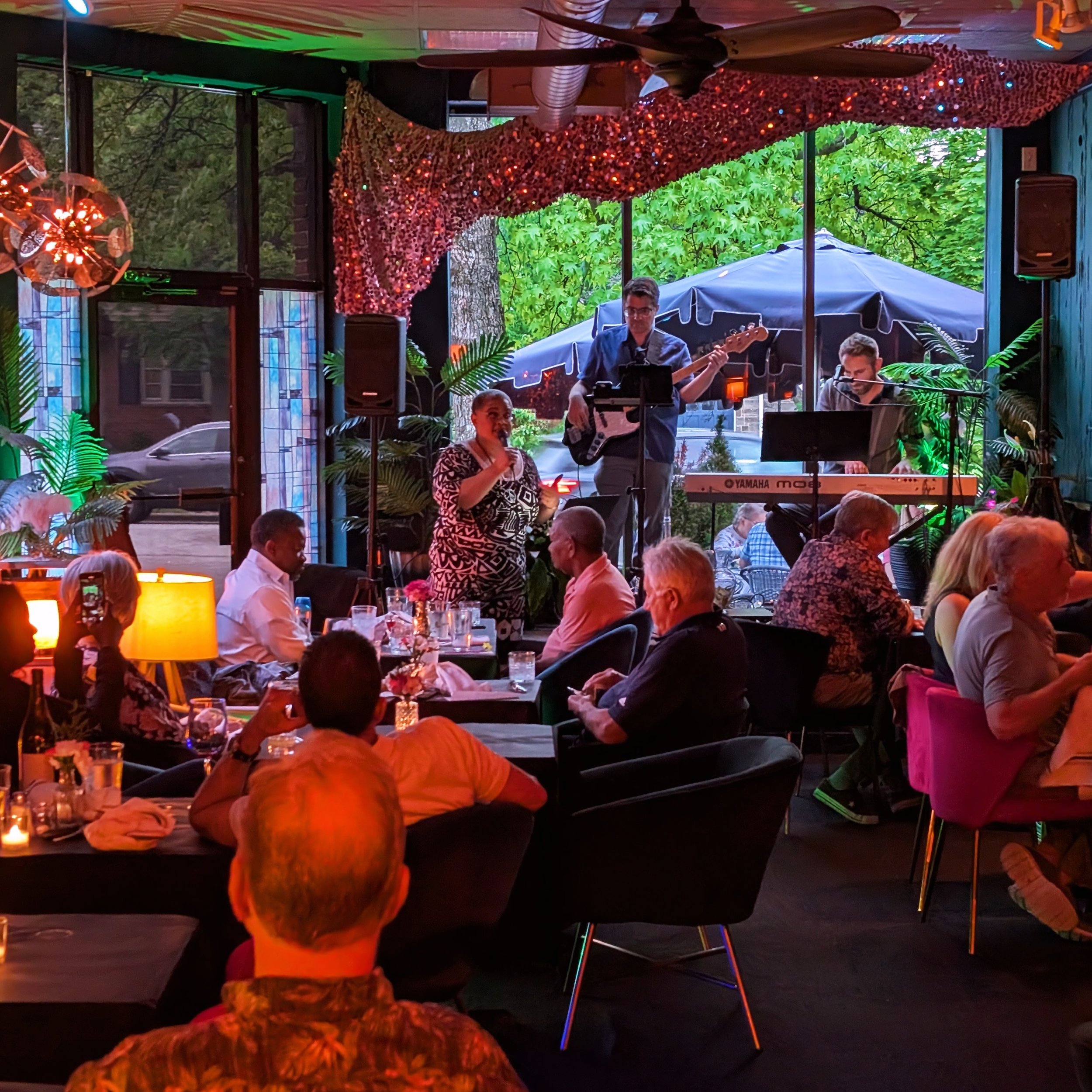 Indoor restaurant with live band performing on stage, guests seated at tables, some taking photos, candles and decorative lighting inside, with greenery visible outside through large windows.