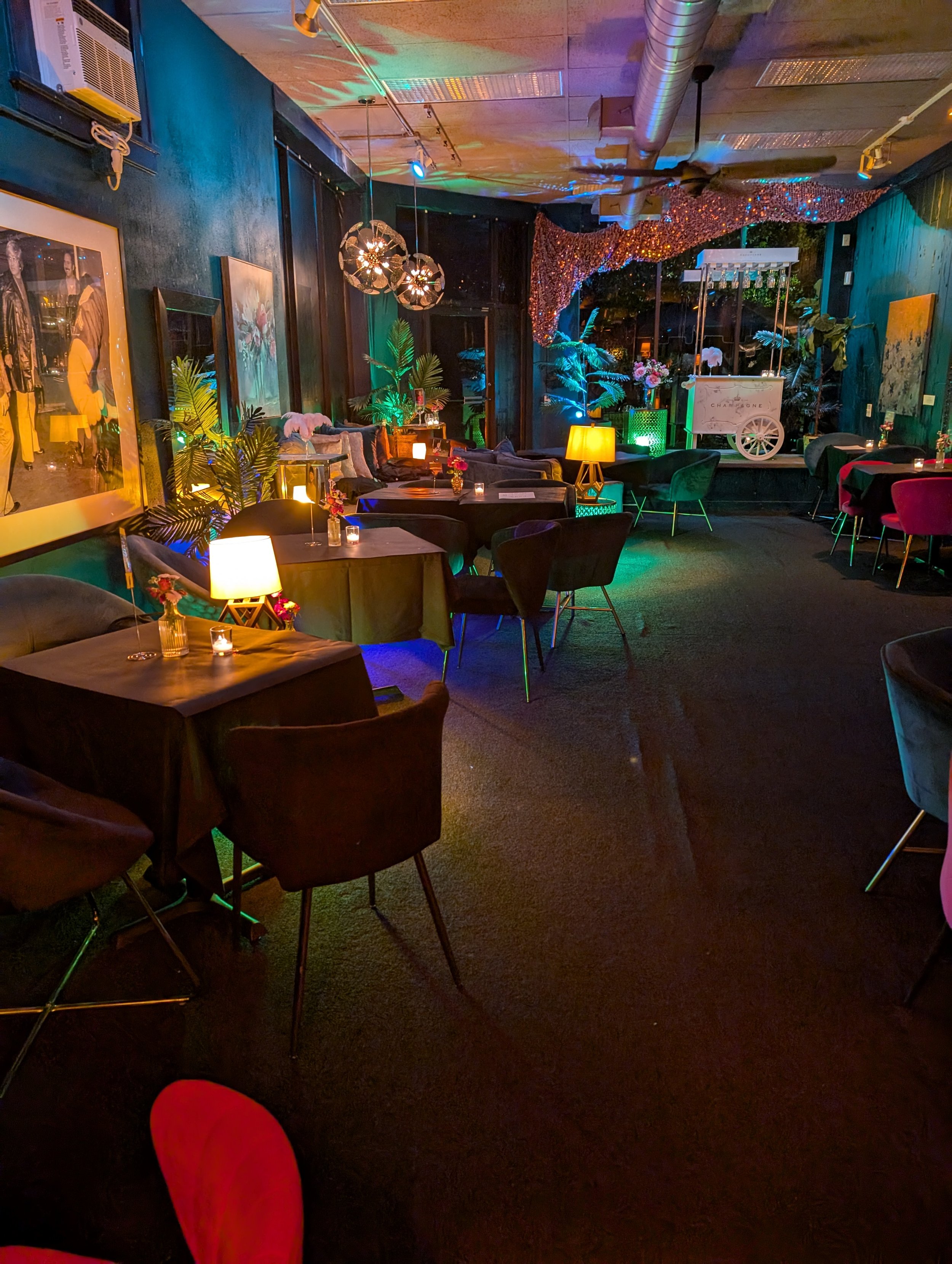 An intimate lounge with dim lighting, decorated with lamps, candles, and plants, and a small stage with a cart in the background.