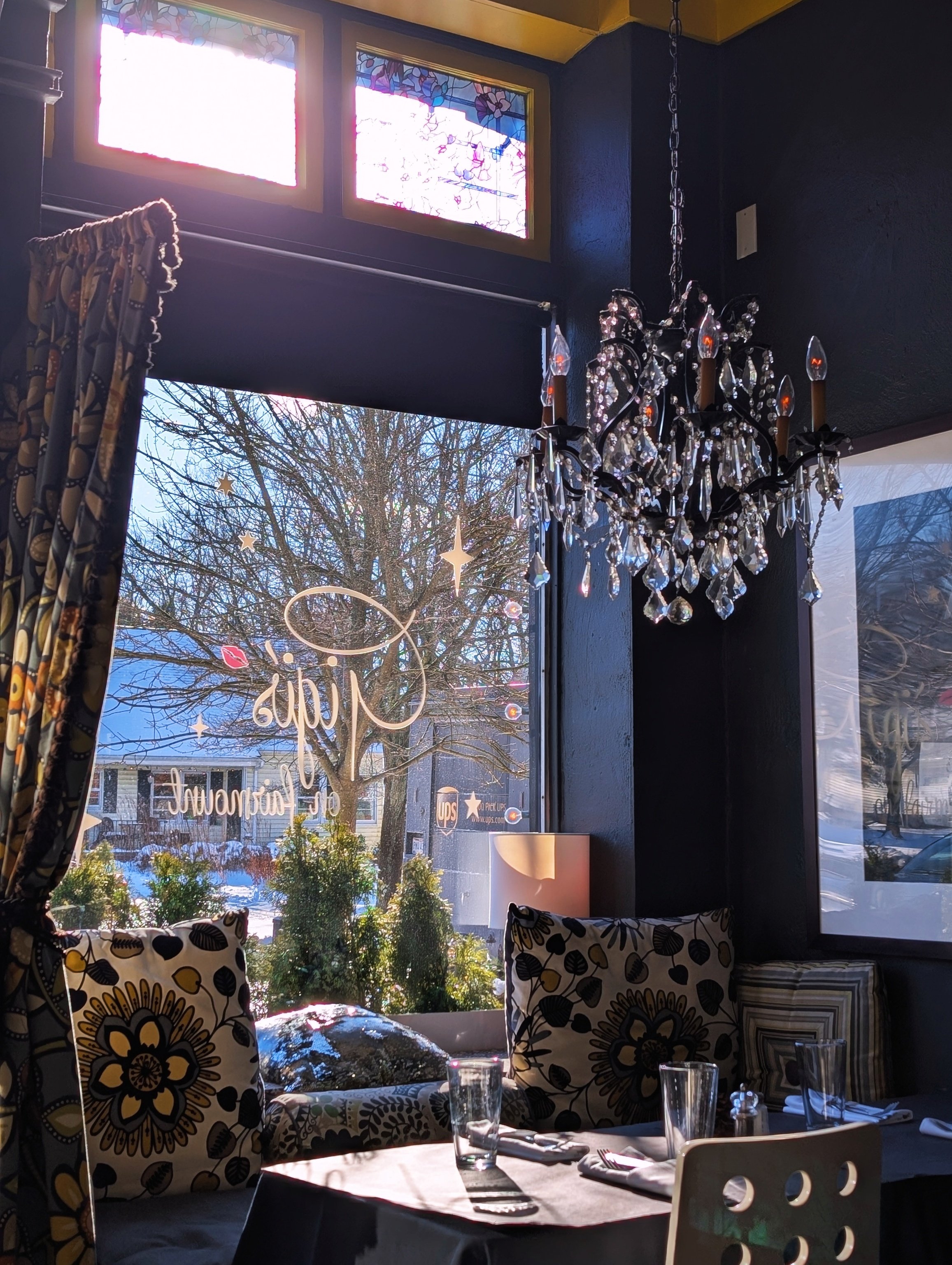 Interior of Gigi's on Fairmount with a chandelier, patterned curtains, and floral cushions, looking out a window with a snowy landscape and leafless trees outside.
