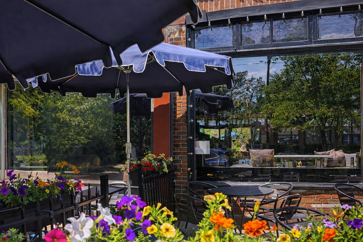 Outdoor patio with tables and chairs, umbrellas, and colorful flowers, reflected in a large window of a brick building.