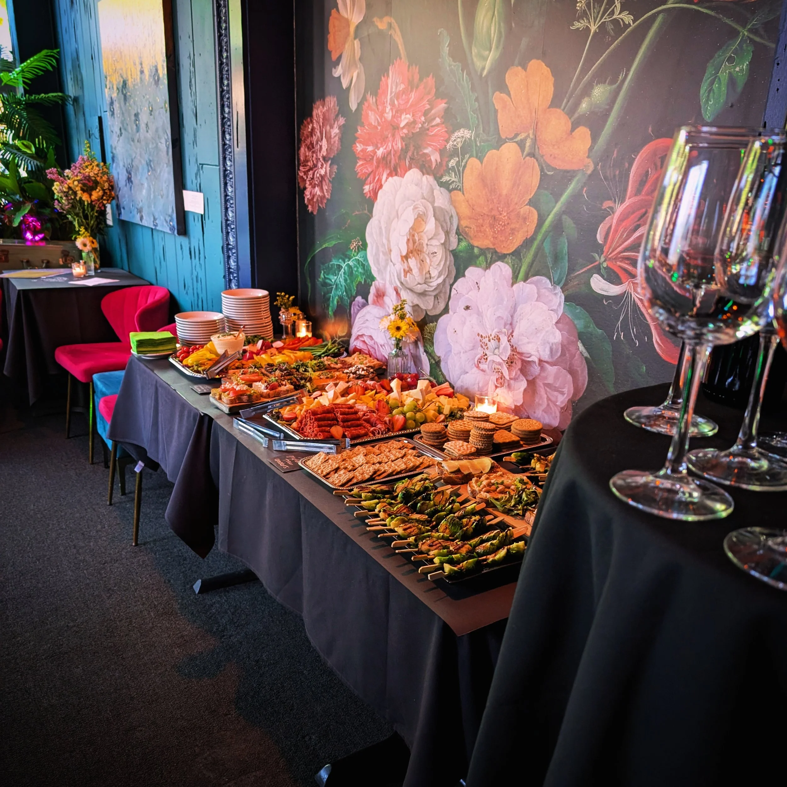 Buffet table with assorted appetizers, cheeses, vegetables, crackers, and skewers, set against a floral mural with pink and white flowers.