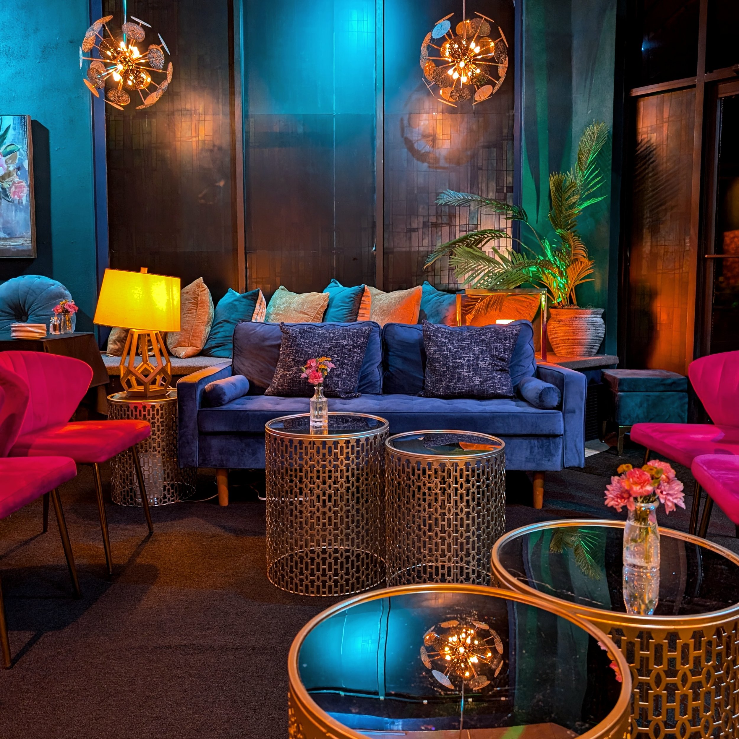 Colorful private space with velvet sofa, pink chairs, gold patterned side tables, decorative lamps, and plants, illuminated with warm and vibrant lighting.