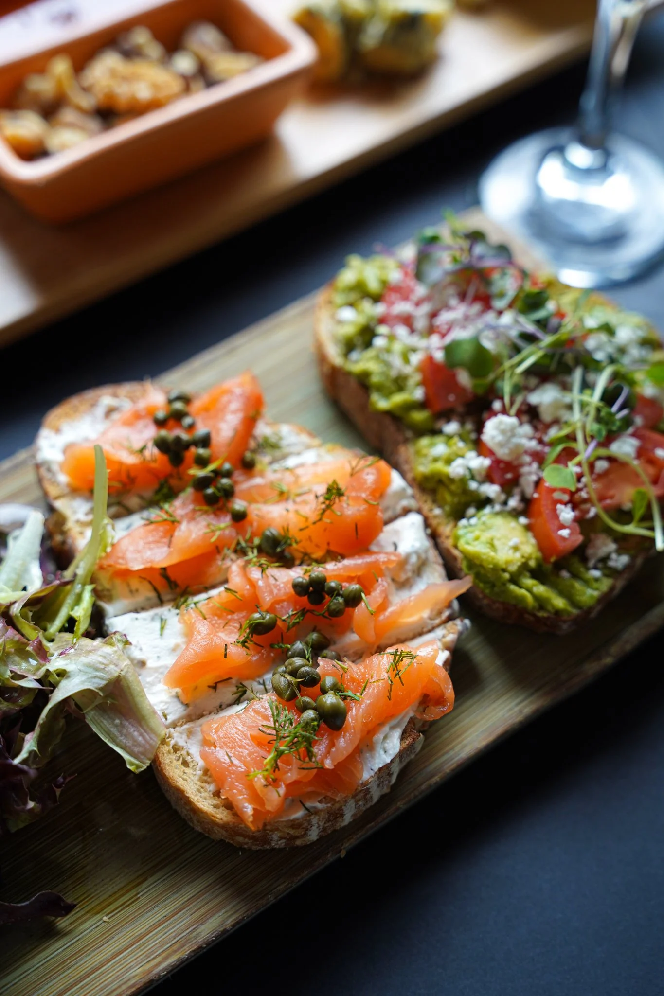 Close-up of two slices of artisan bread topped with spread, smoked salmon, capers, and dill, served on a wooden platter with salad greens on the side.