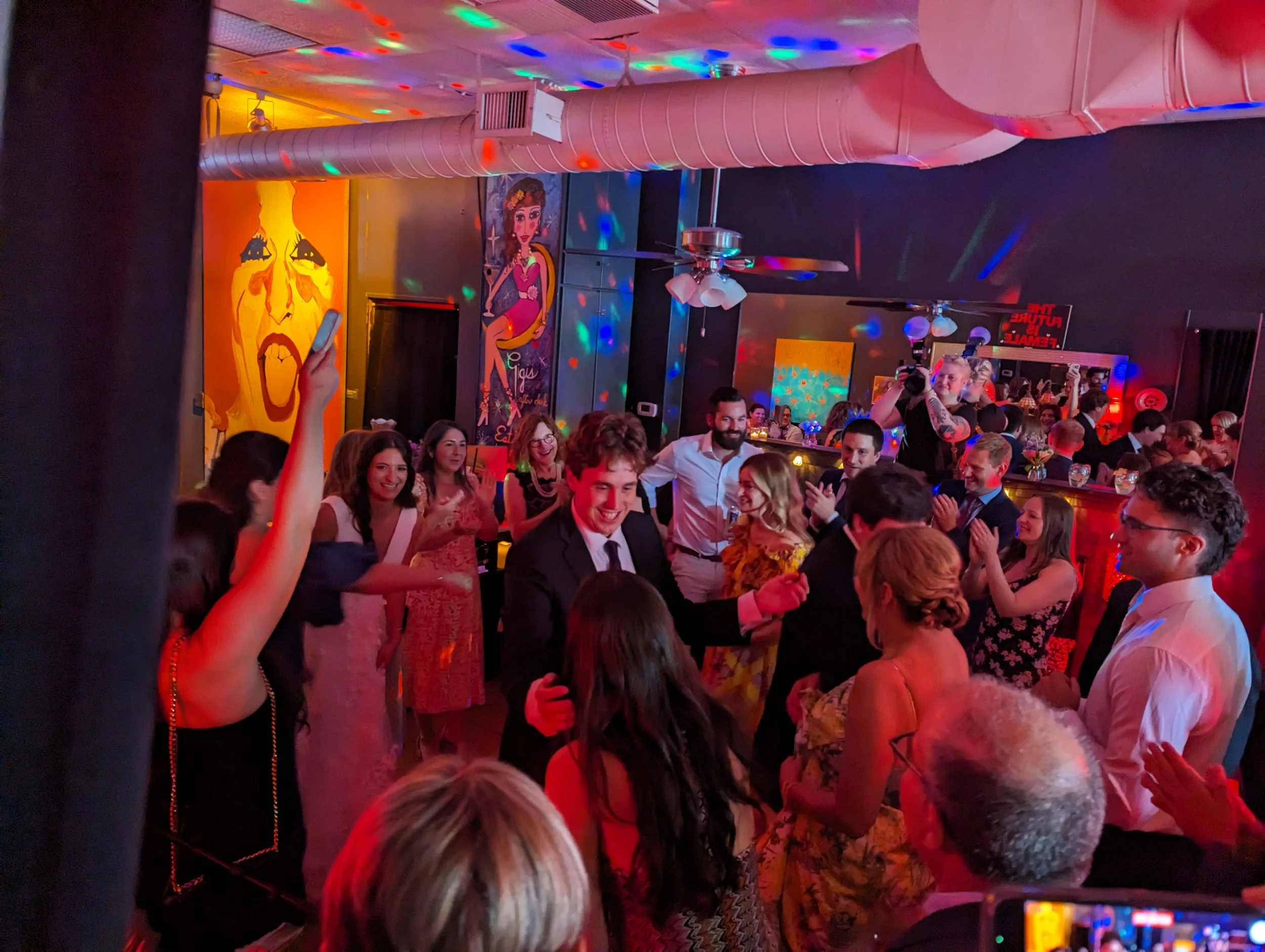 People dance and celebrate at a party in a colorful, decorated venue with artwork and vibrant lighting.