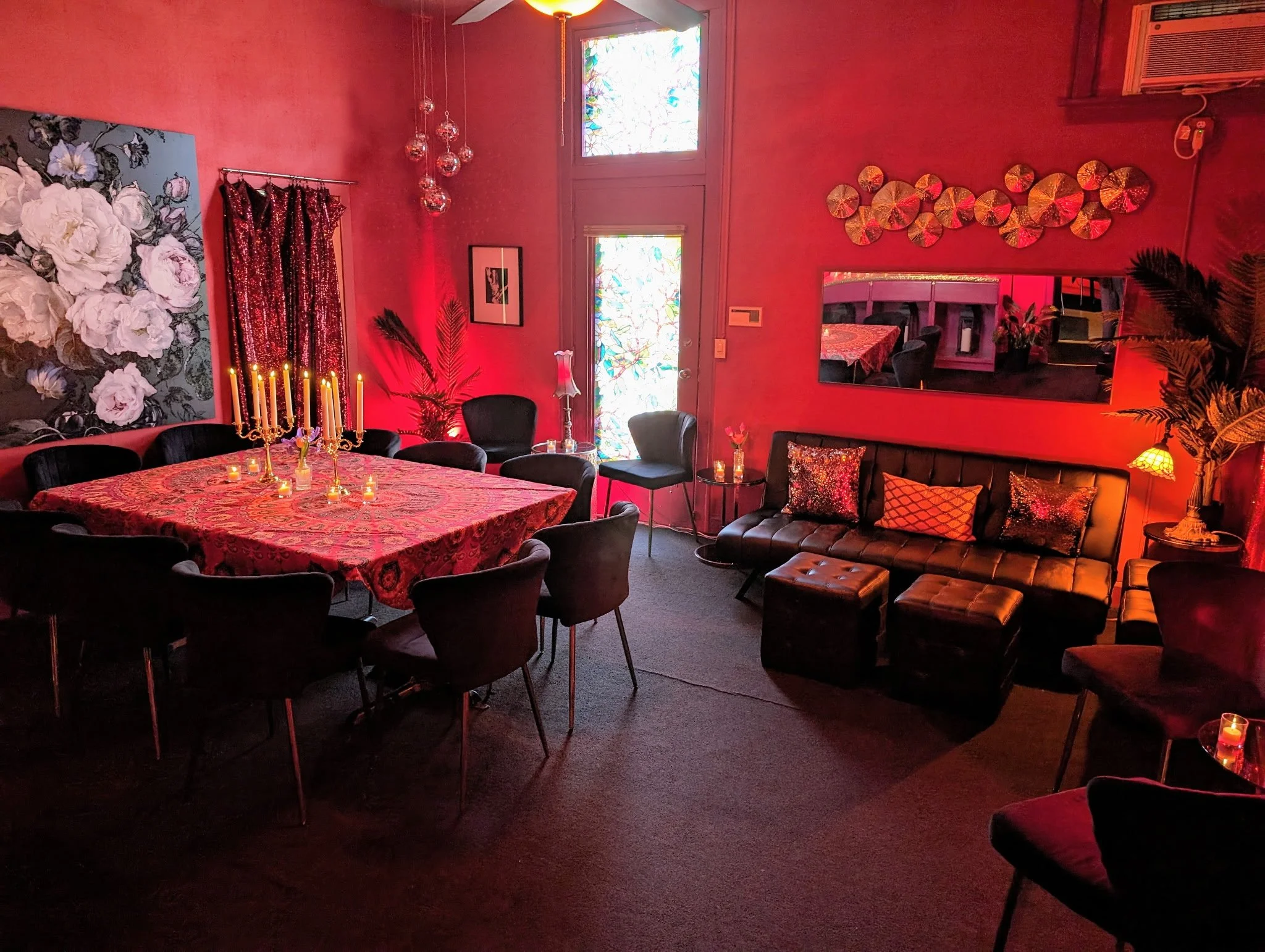 A cozy dining and lounge area with red ambient lighting, a round table with a red patterned tablecloth, surrounded by black chairs, a black leather couch with decorative pillows, multiple decorative wall art pieces, and warm lighting fixtures.
