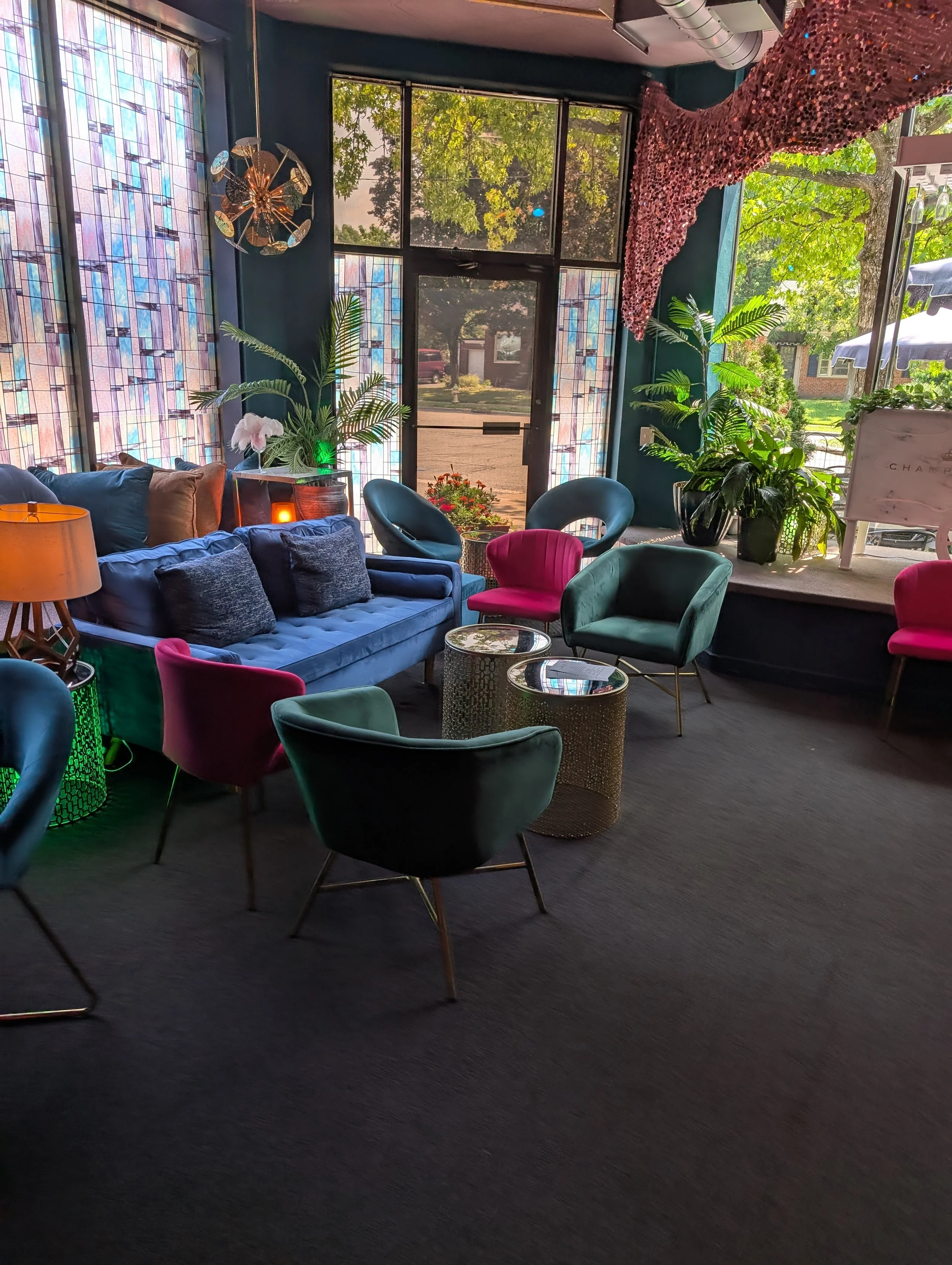 Colorful lounge area with green, pink, and blue chairs around gold metallic tables, a blue velvet sofa with pillows, potted plants, stained glass windows, and a pink sequined curtain in a cozy interior.