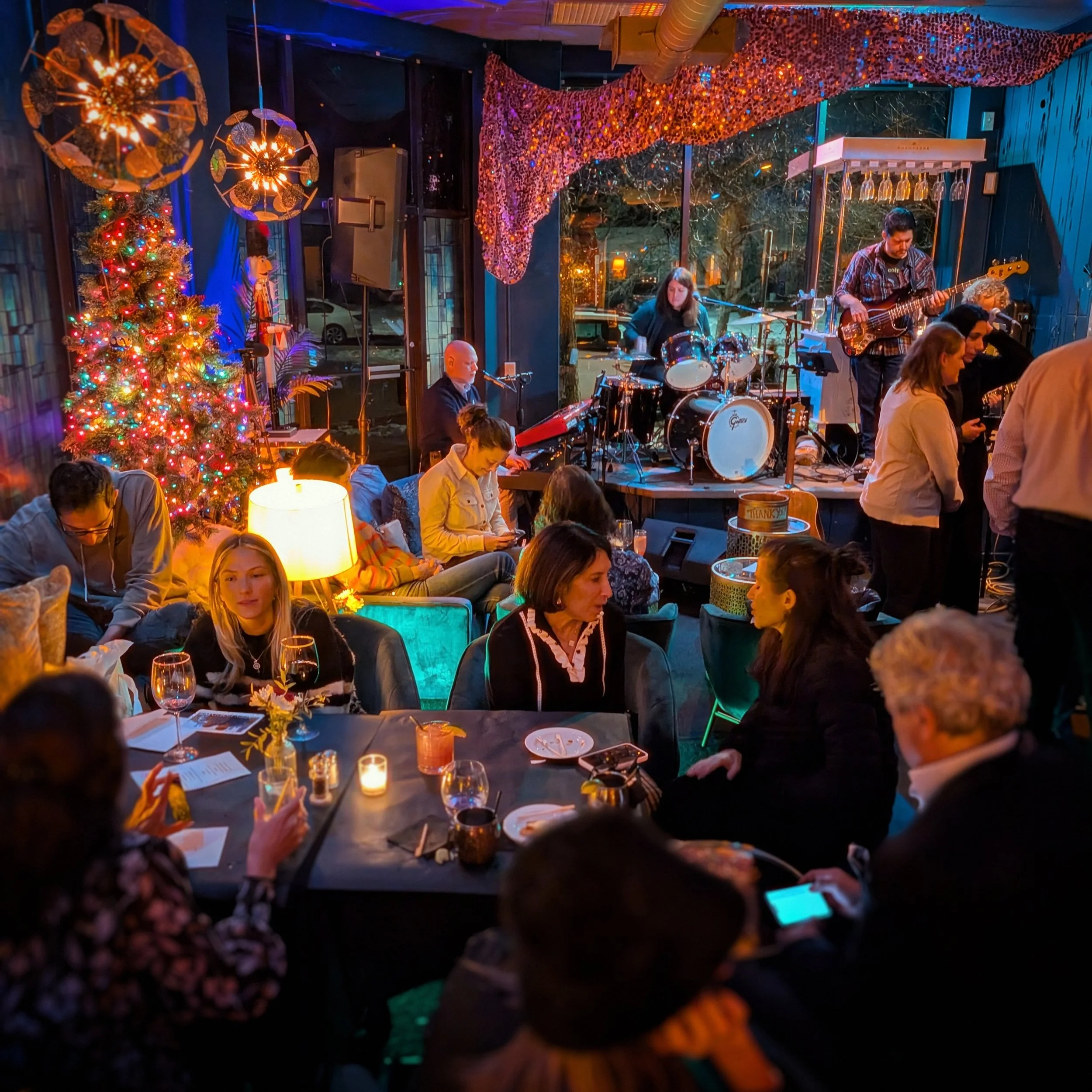 People gathered in a cozy restaurant with christmas decorations, a Christmas tree, and a live band performing on stage.