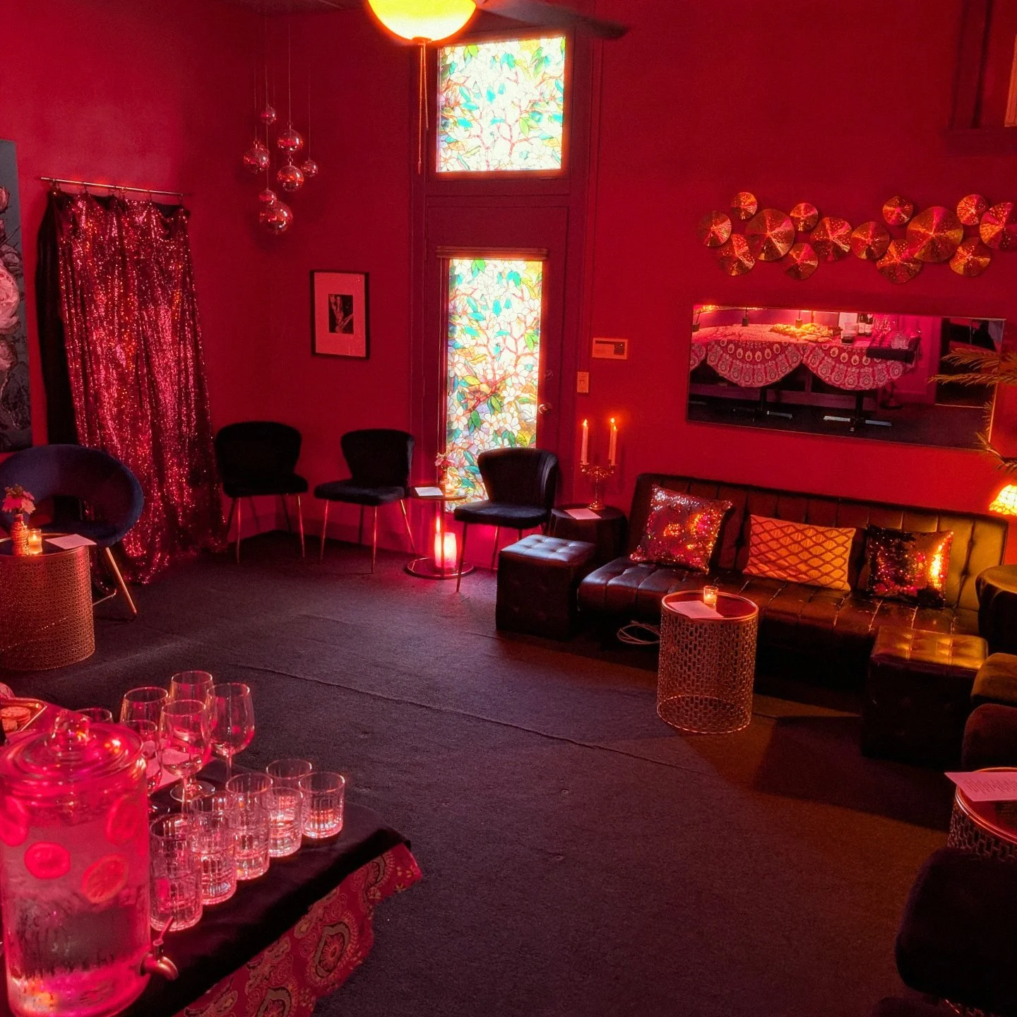 A cozy, dimly lit living room decorated with red lighting. It features black chairs, a black leather sofa with colorful pillows, a small round table with a lit candle, and a table with glasses and a water dispenser. The walls have decorative plates, 