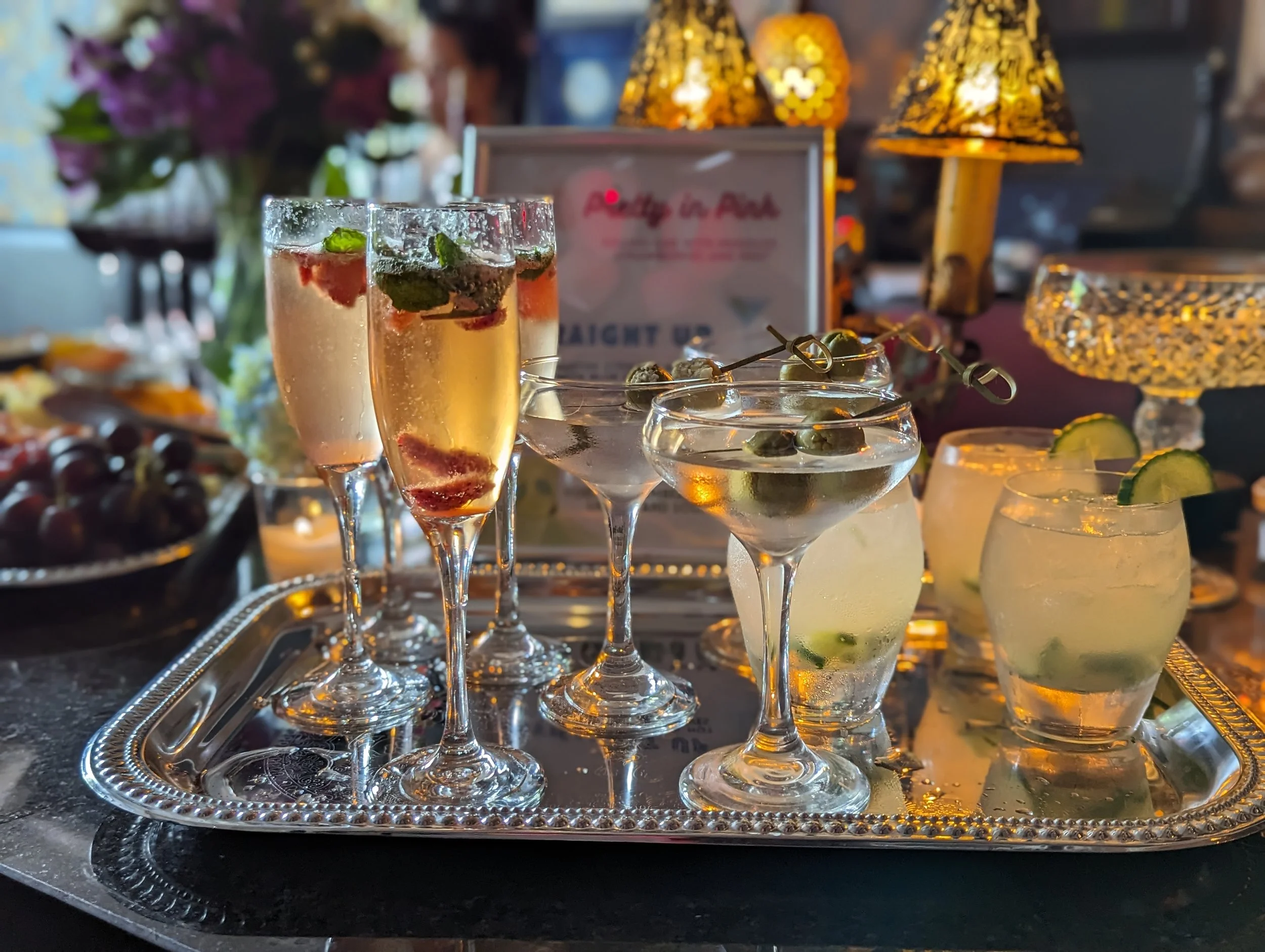 Tray of assorted cocktails, including champagne flutes with strawberries and mint, martinis with olives, and glasses with lime slices, set on a silver tray with a blurred background featuring lamps and flowers.
