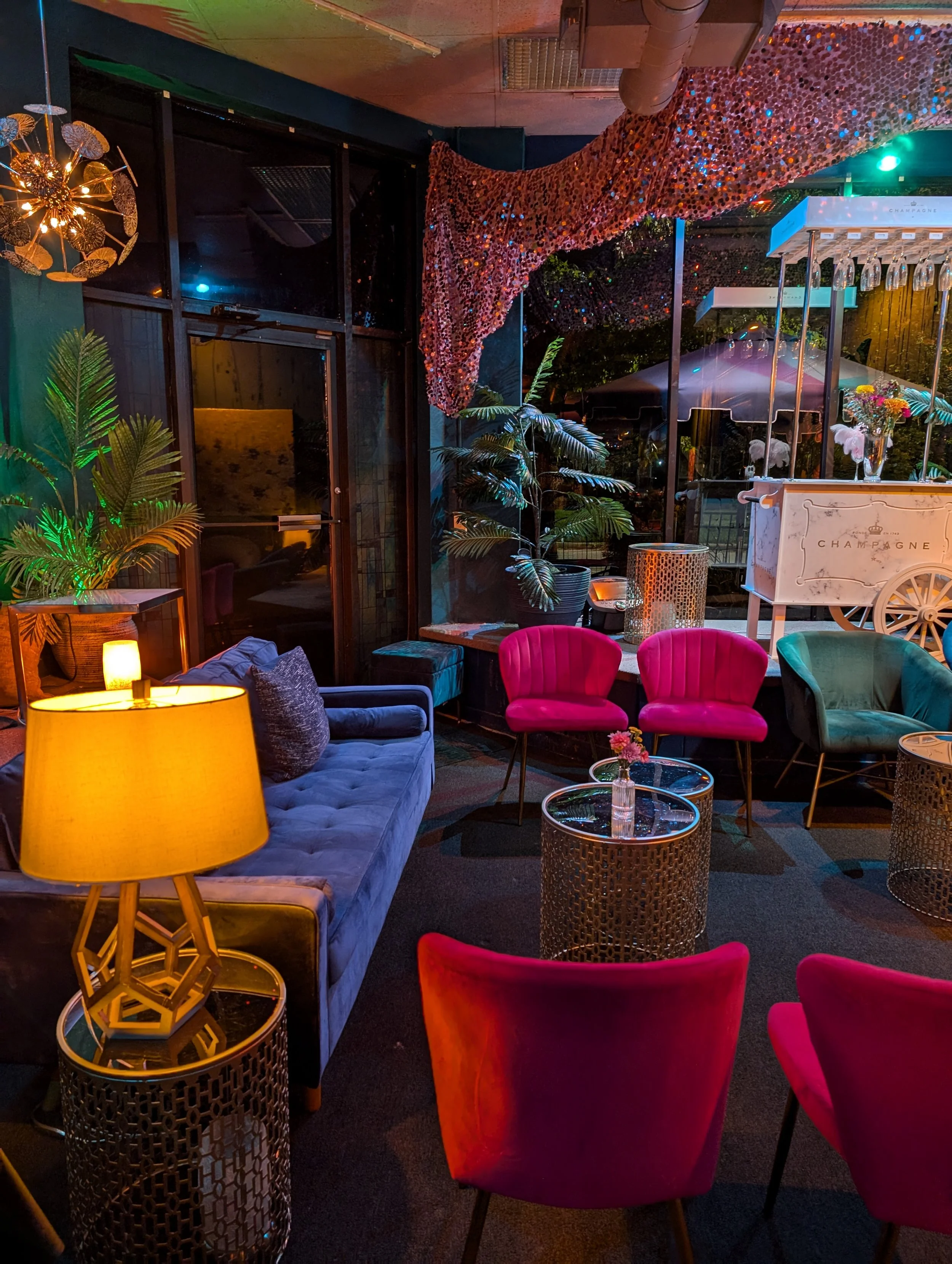 Interior of a stylish lounge or restaurant with colorful furniture including pink and teal chairs, a blue velvet sofa, and glass-topped tables with small flower vases. Warm and colorful lighting accents the space, with decorative plants, a lamp, and 