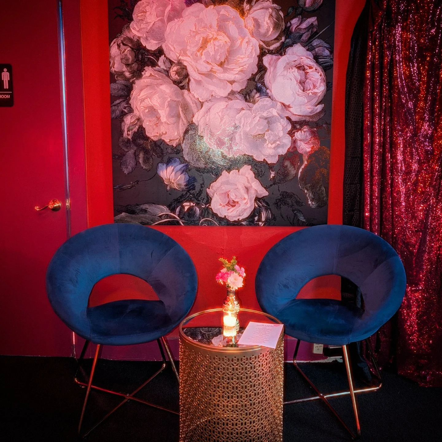 A cozy corner with two blue velvet chairs facing each other, a small round gold table in between holding a small flower arrangement and a candle, with a large floral painting of pink flowers on a black background on the red wall behind, and a red seq
